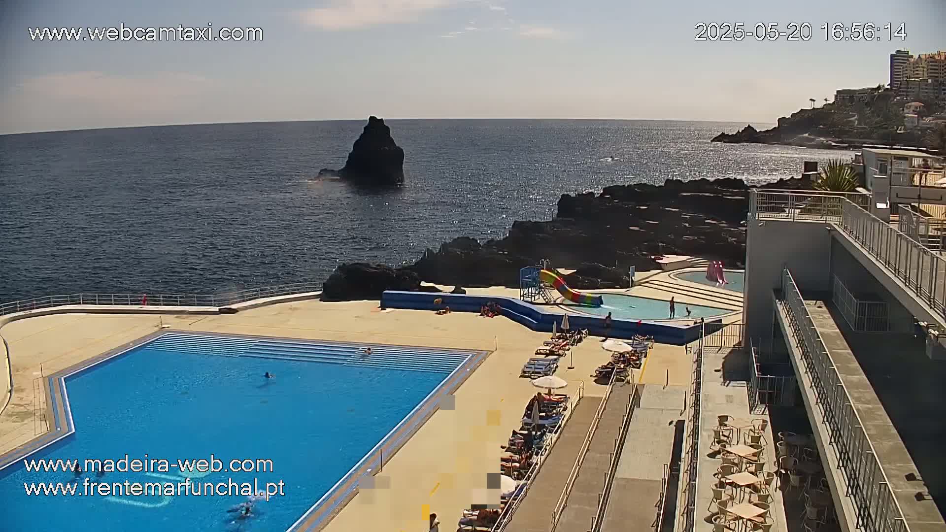 An outdoor pool complex with a large swimming pool, a smaller pool with a waterslide, and sunbathers is situated on a rocky coast overlooking a calm ocean under a sunny sky.
