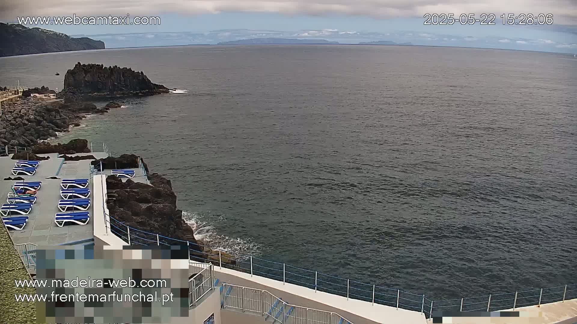 A rocky coastline with a sun deck featuring several empty lounge chairs overlooks a calm, dark-grey ocean under a cloudy sky.