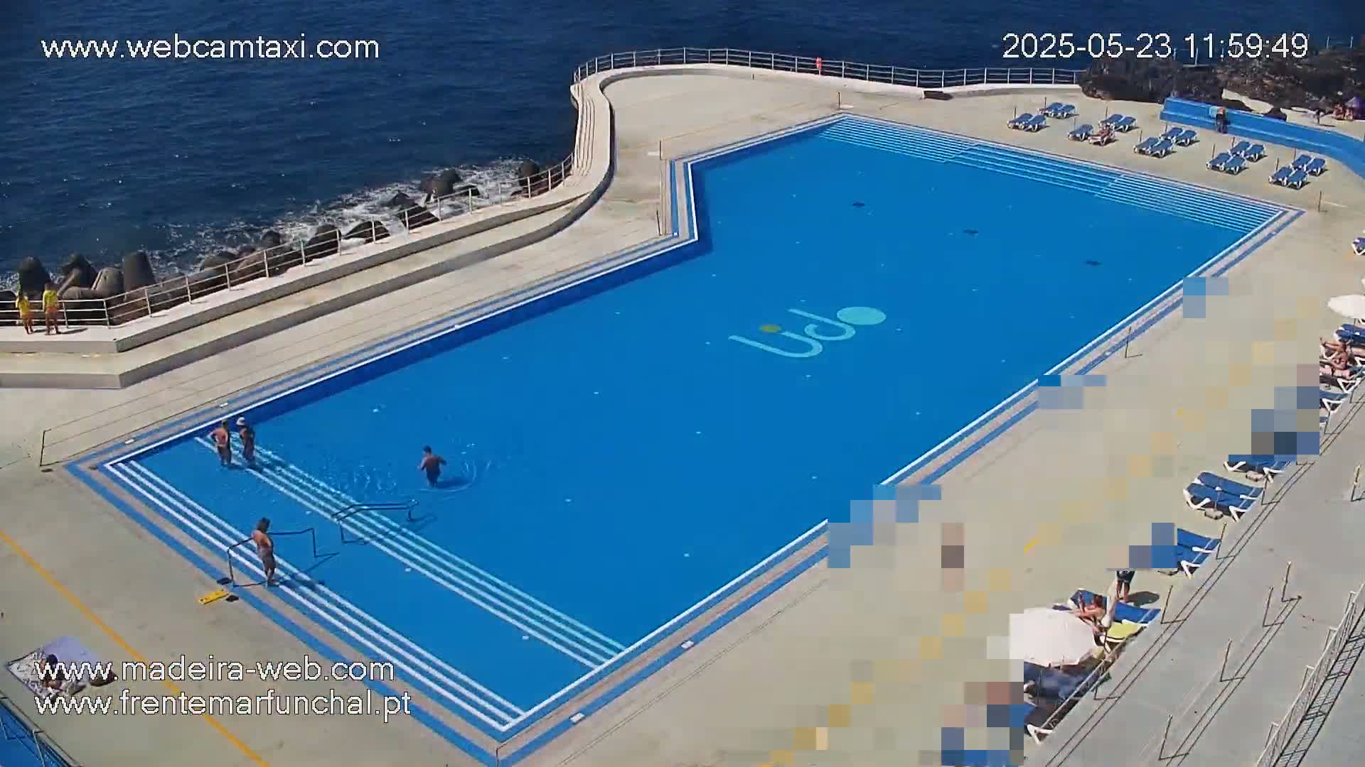 An outdoor pool with several people swimming and sunbathing on a sunny day next to a rocky ocean shore.