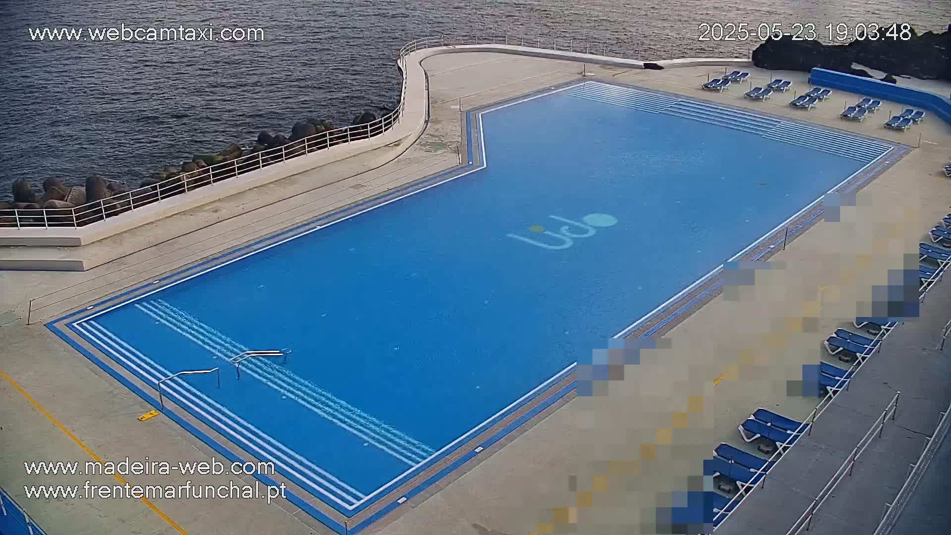An outdoor, blue rectangular swimming pool with several lounge chairs is situated next to a body of dark water on a mostly sunny day.