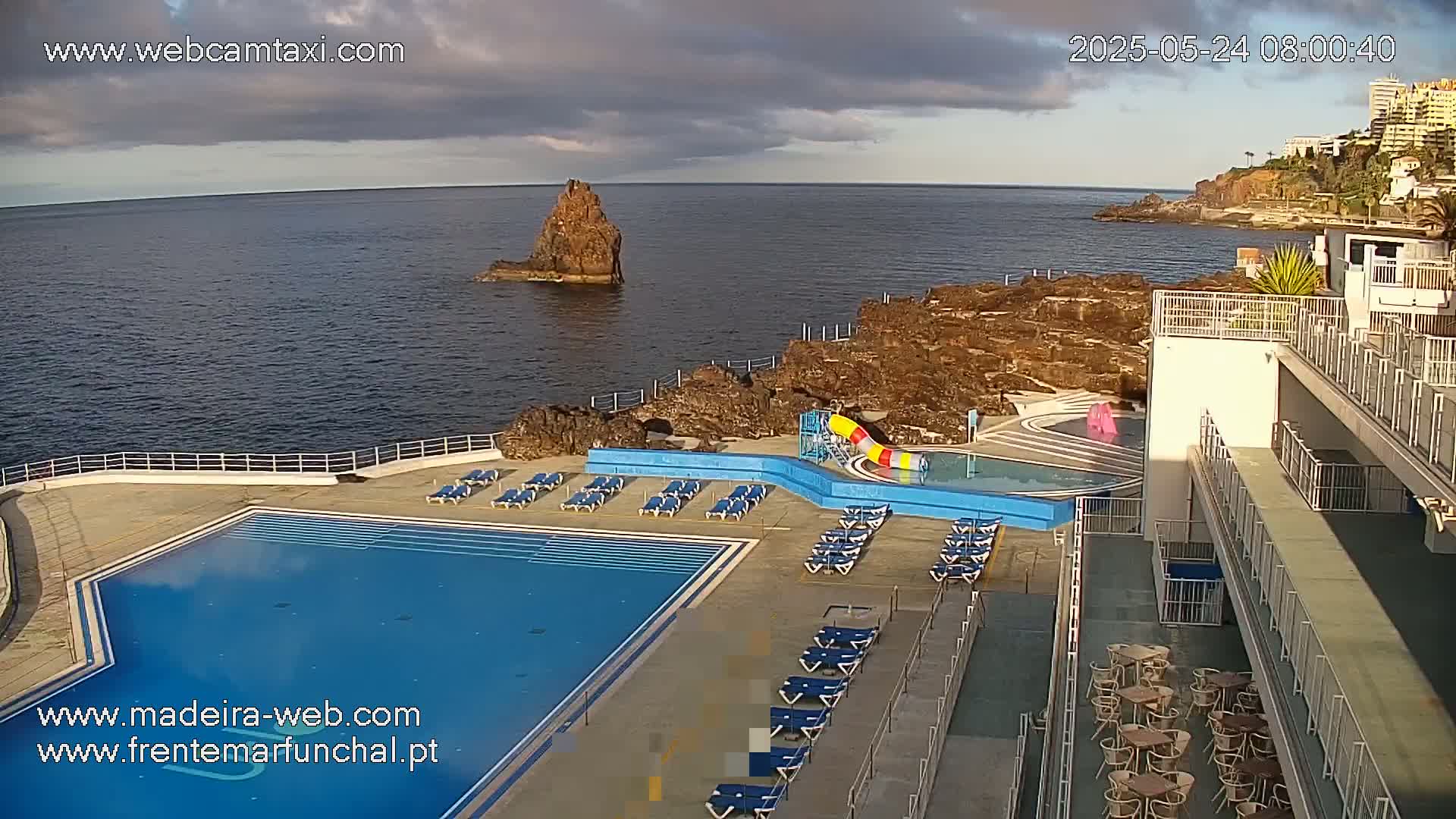 An outdoor pool area with lounge chairs and a water slide overlooks a rocky coastline and ocean under partly cloudy skies.