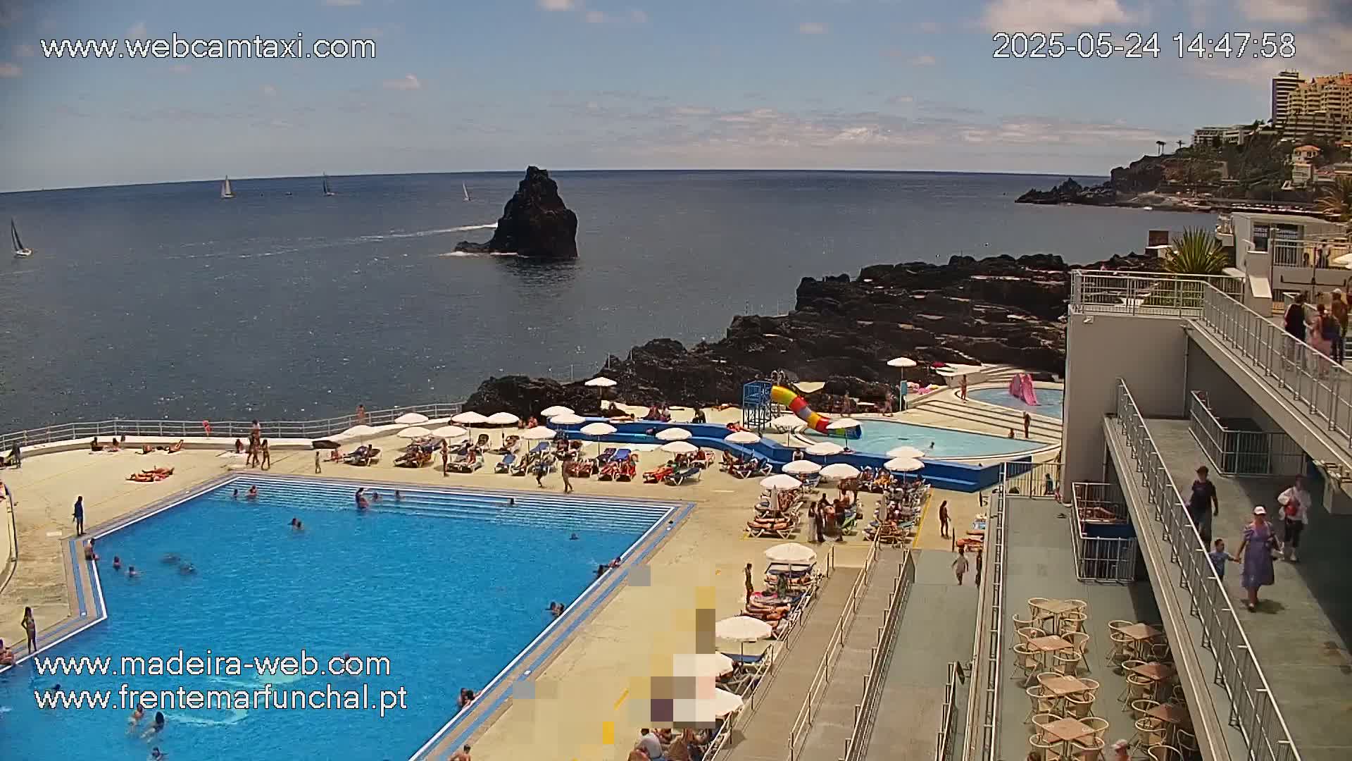 An outdoor swimming pool complex is situated on a rocky coastline overlooking a calm ocean on a sunny day, with people swimming and relaxing around the pool and on the surrounding decks.