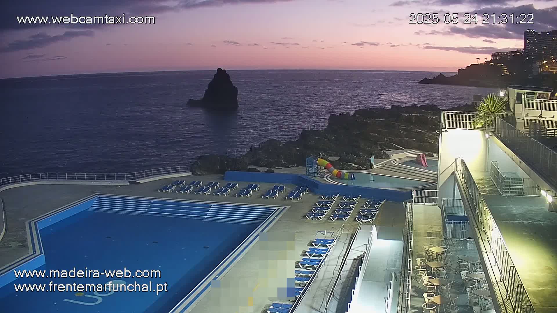 An outdoor swimming pool area with lounge chairs sits on a cliff overlooking a dark ocean at twilight.