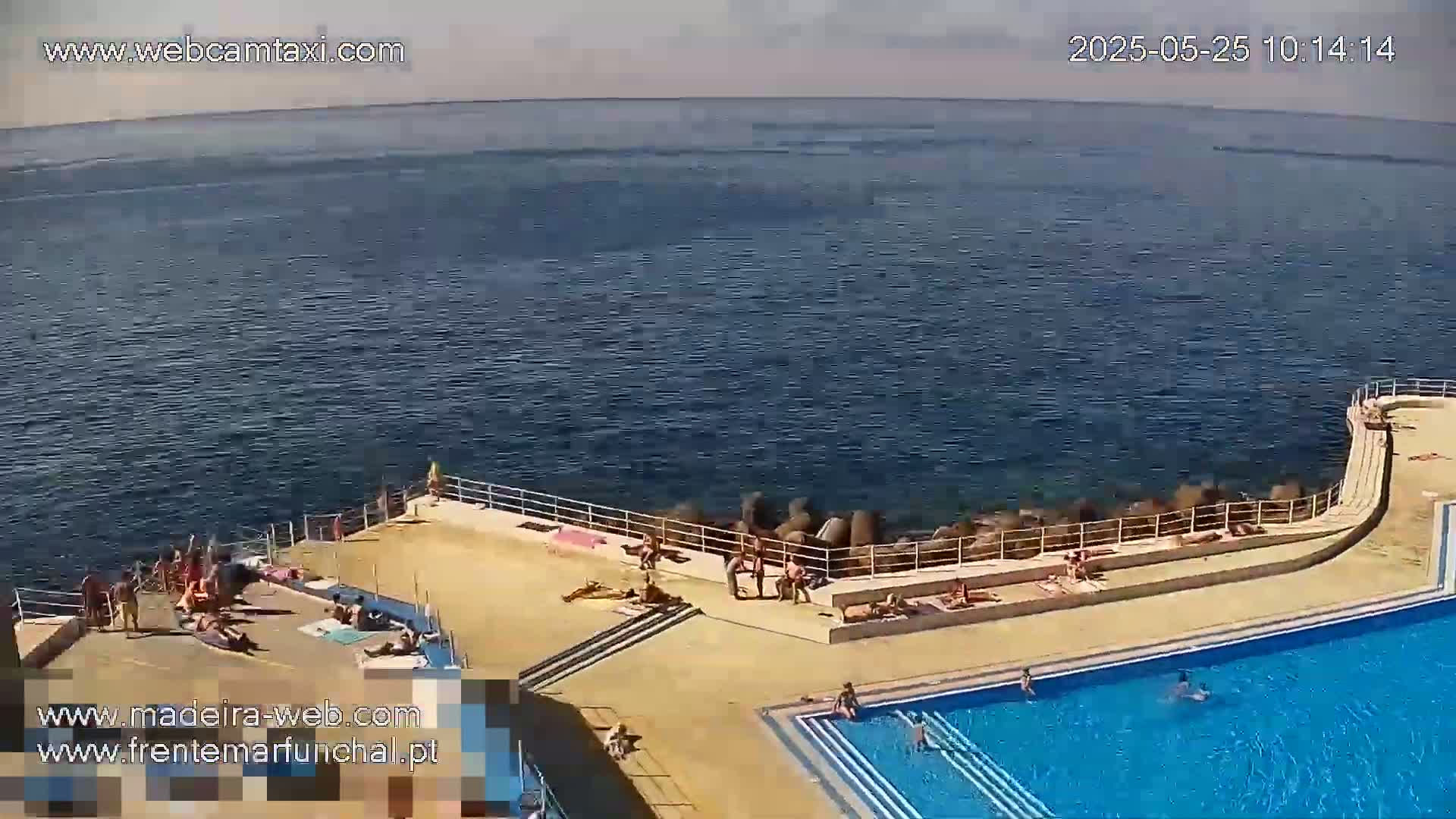 An outdoor swimming pool complex next to the ocean on a sunny day, with people sunbathing and swimming.
