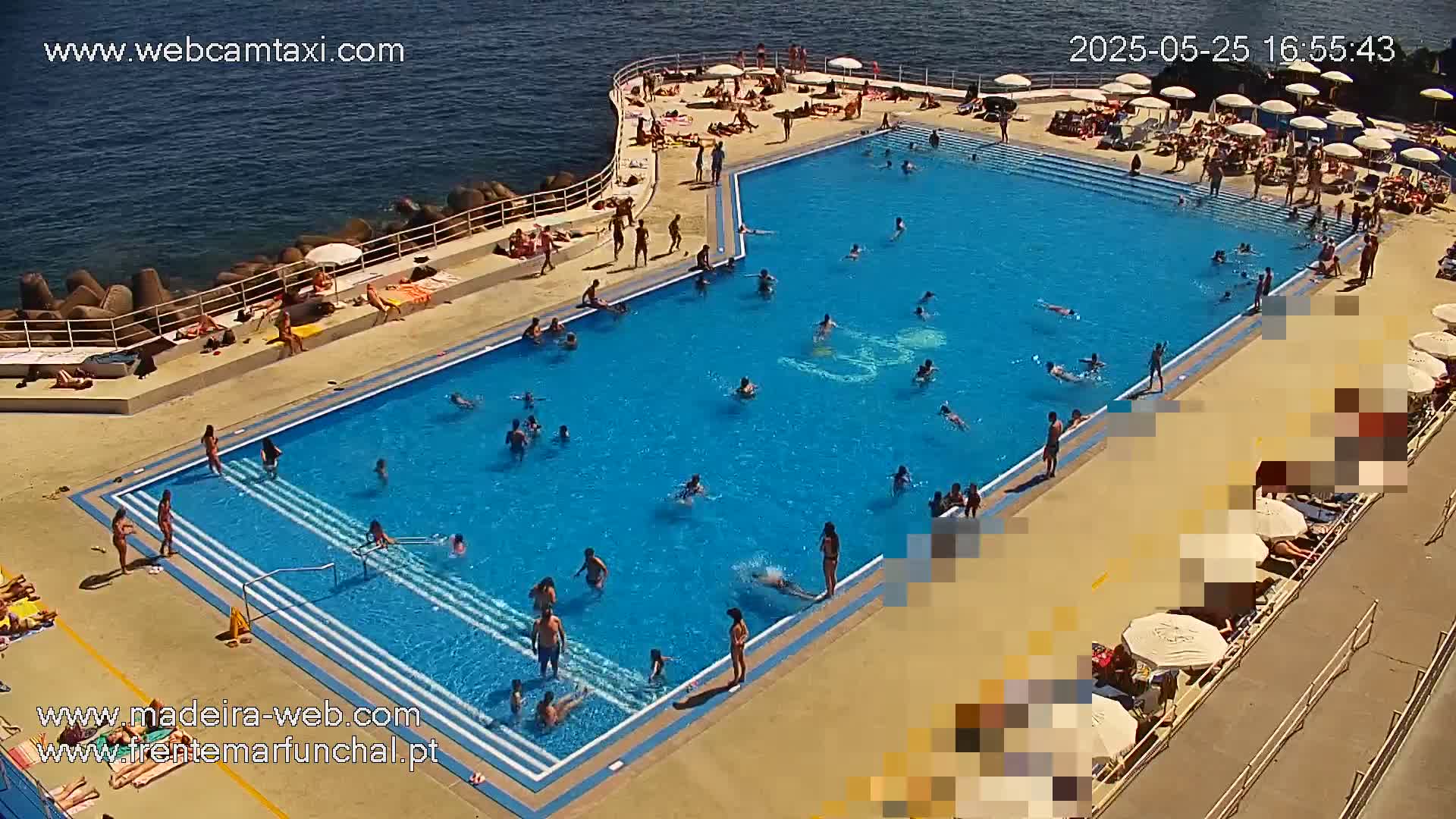 A large outdoor swimming pool filled with many people on a sunny day next to a rocky ocean shore.