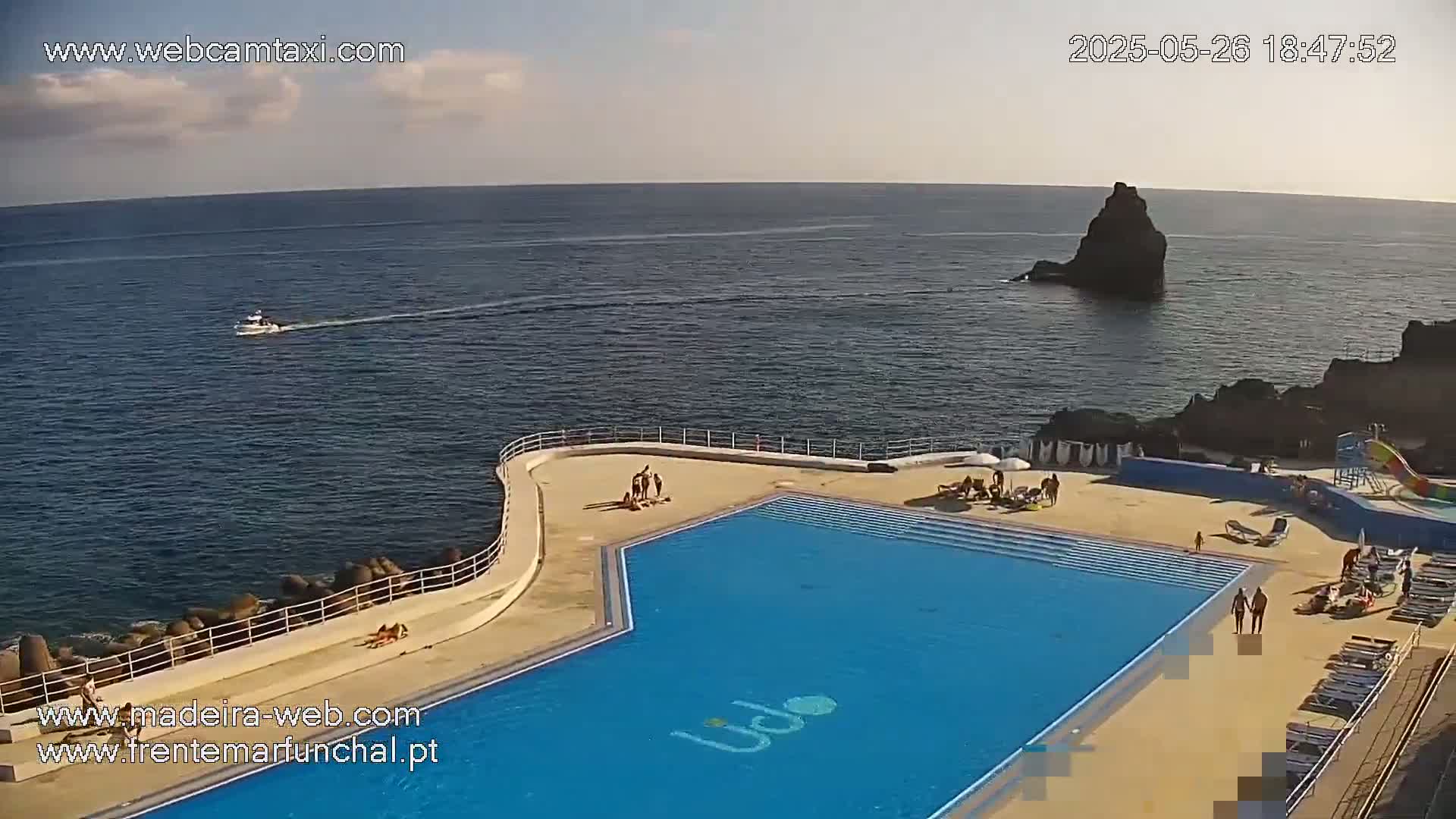 An outdoor swimming pool with people sunbathing and swimming is situated on a cliff overlooking a calm ocean with a small boat and a dark rock formation visible in the distance under a partly cloudy sky.