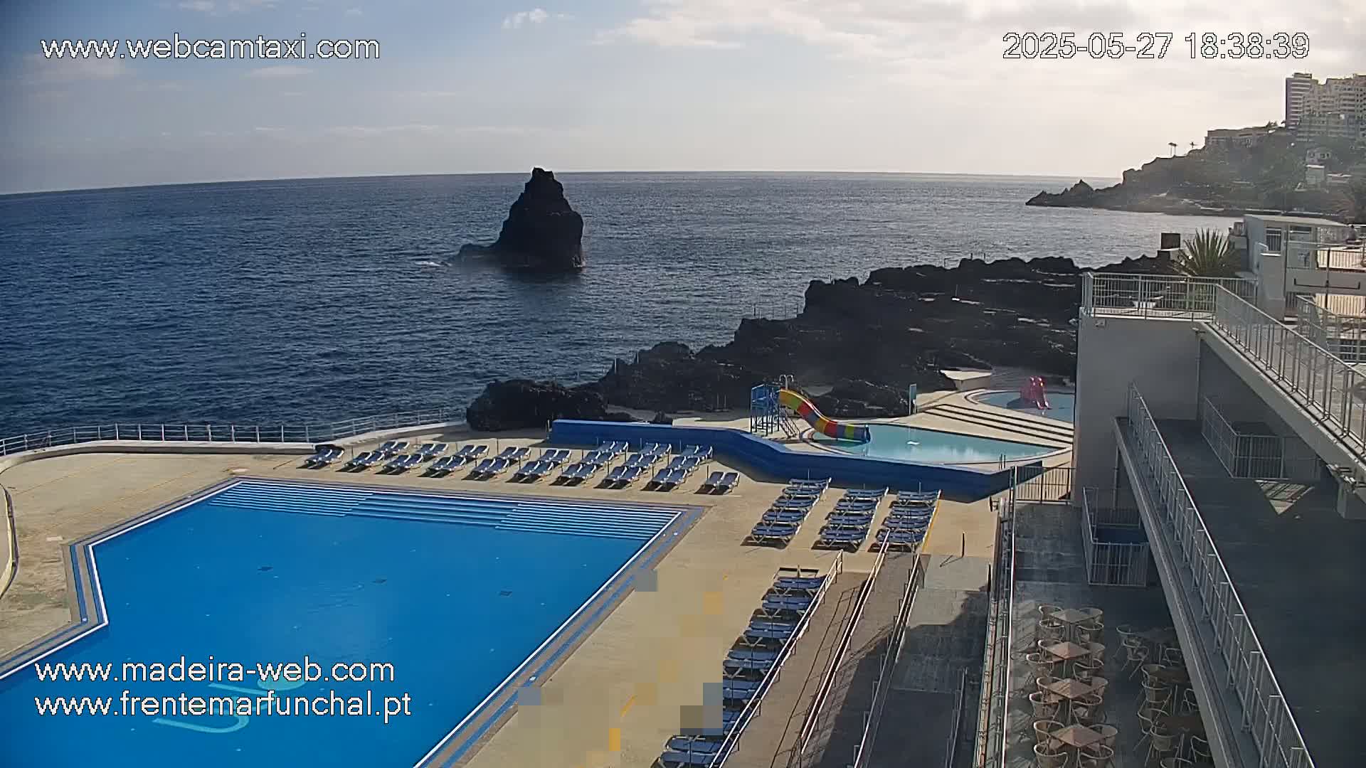 An outdoor pool and lounge area next to a rocky ocean coast on a partly sunny day.