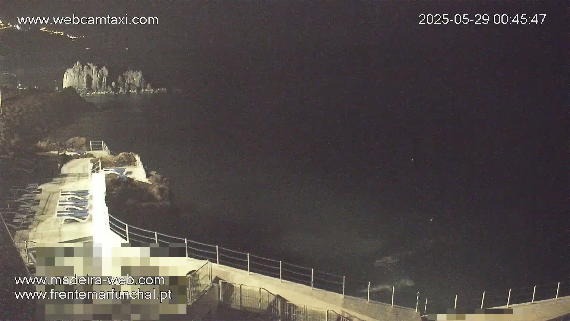 A nighttime view of a dark ocean, with a walkway and lounge chairs along a rocky cliffside.