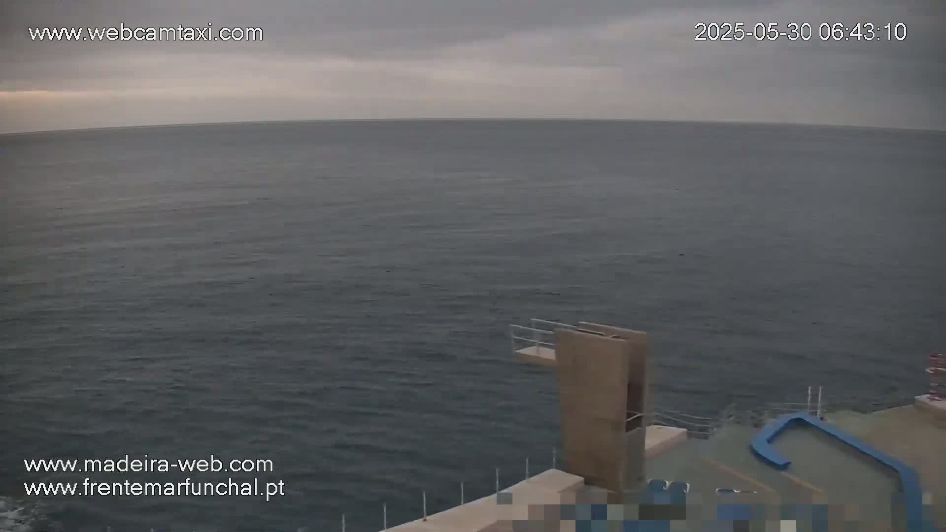 A high-angle view shows a calm, dark-grey ocean under an overcast sky, with a diving platform and other structures on a nearby shore.