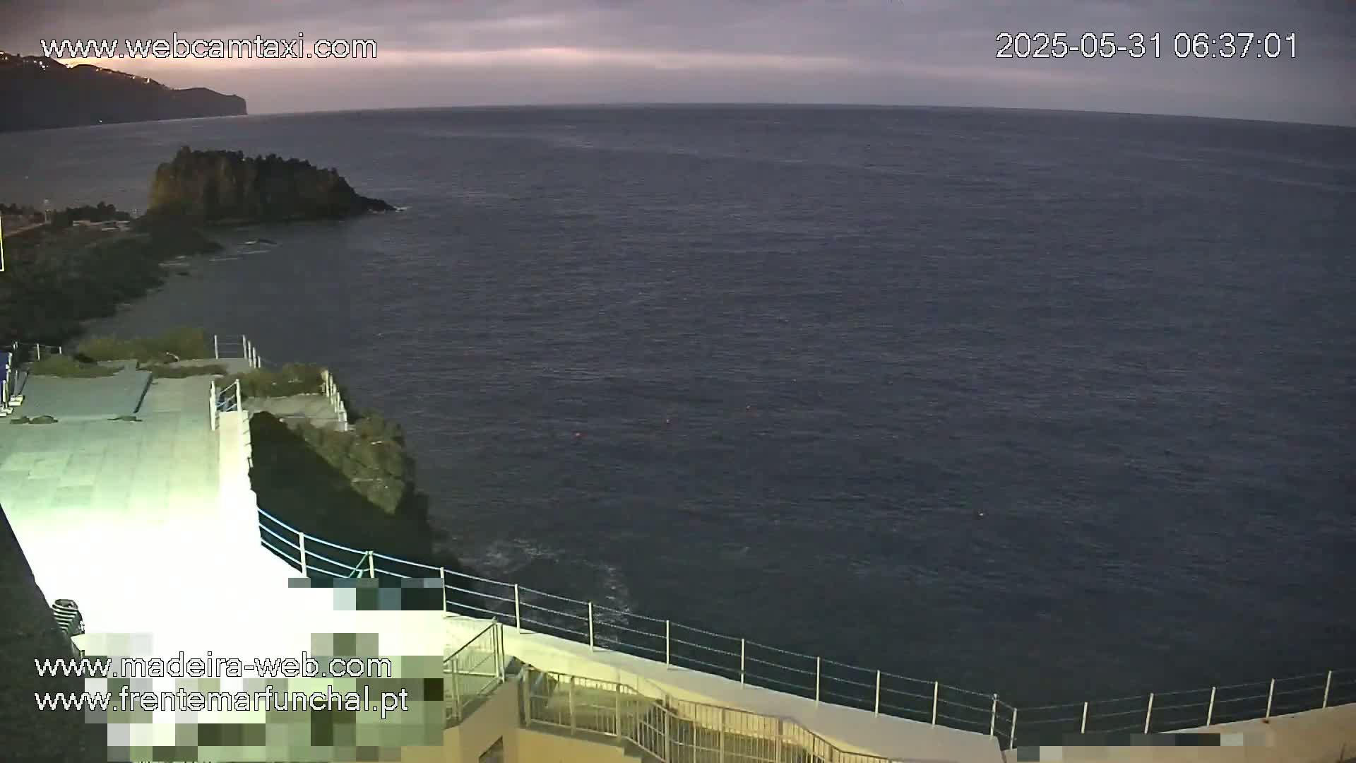 A dark, calm ocean stretches to a distant shoreline under an overcast sky, viewed from a high vantage point overlooking a paved area with a railing.