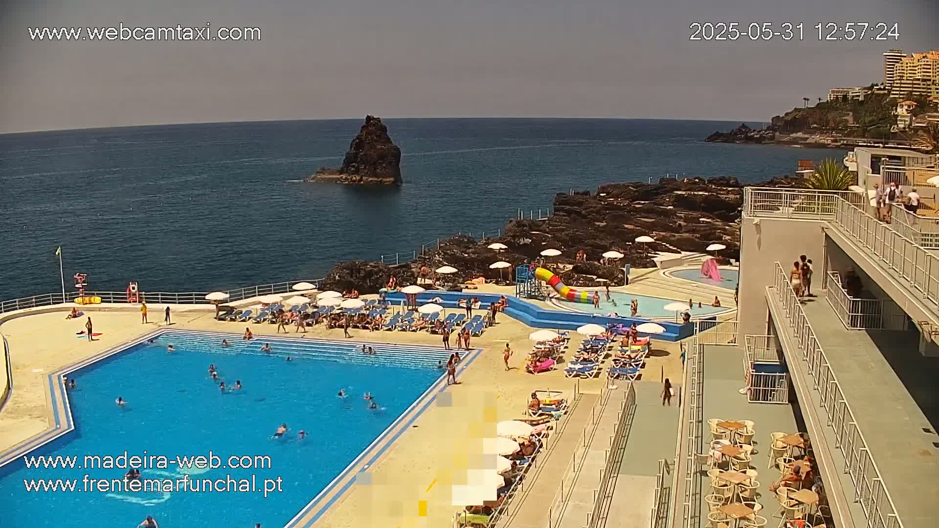 An outdoor pool complex with many people swimming and sunbathing next to the ocean on a sunny day.