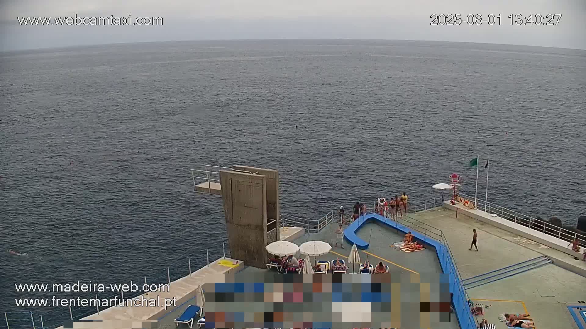 An overcast day at an outdoor swimming area shows a mostly calm body of water, a diving platform, and people lounging on the deck and in the water.