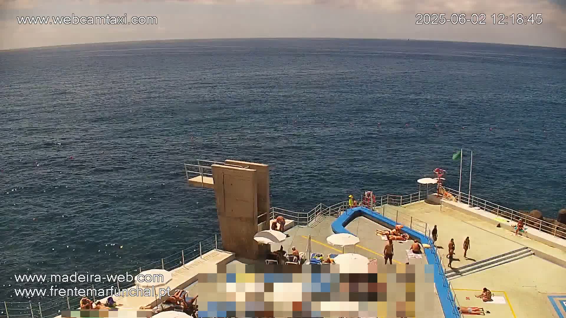 An outdoor swimming area with a diving platform overlooks a calm, blue ocean under a partly cloudy sky.