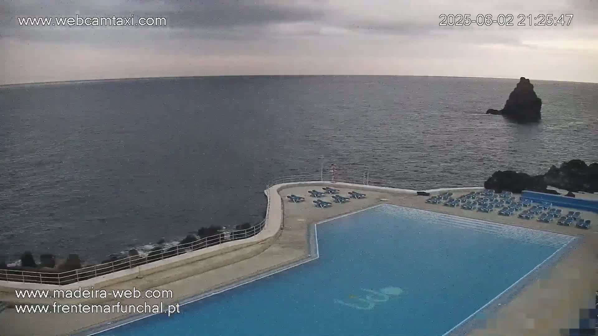 An outdoor infinity pool with lounge chairs sits on a cliff overlooking a dark, calm ocean under an overcast sky.