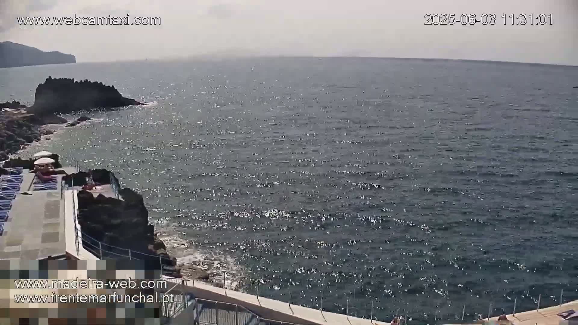 A mostly sunny day shows a rocky coast with a small pool area overlooking a dark blue, somewhat choppy ocean.