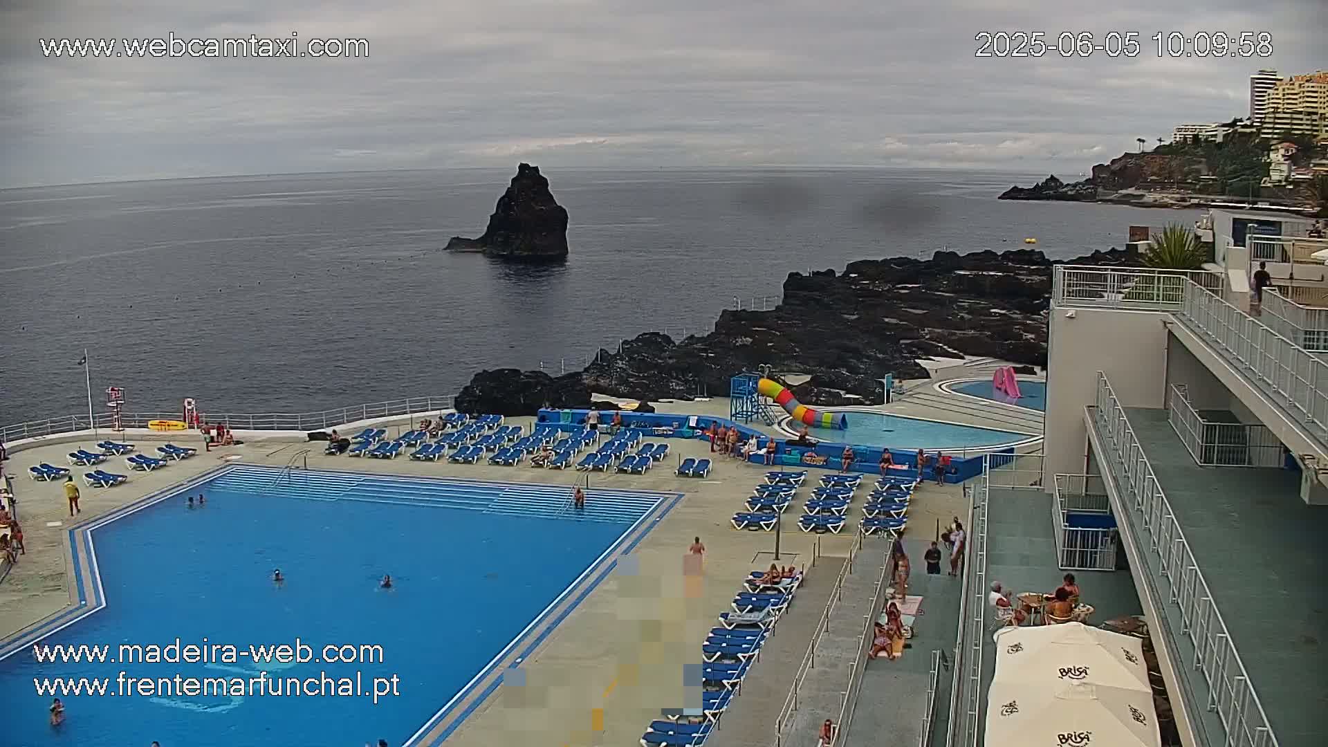 An outdoor pool area with many lounge chairs and a water slide overlooks a dark-colored ocean, with a rocky outcrop visible in the distance under a cloudy sky.