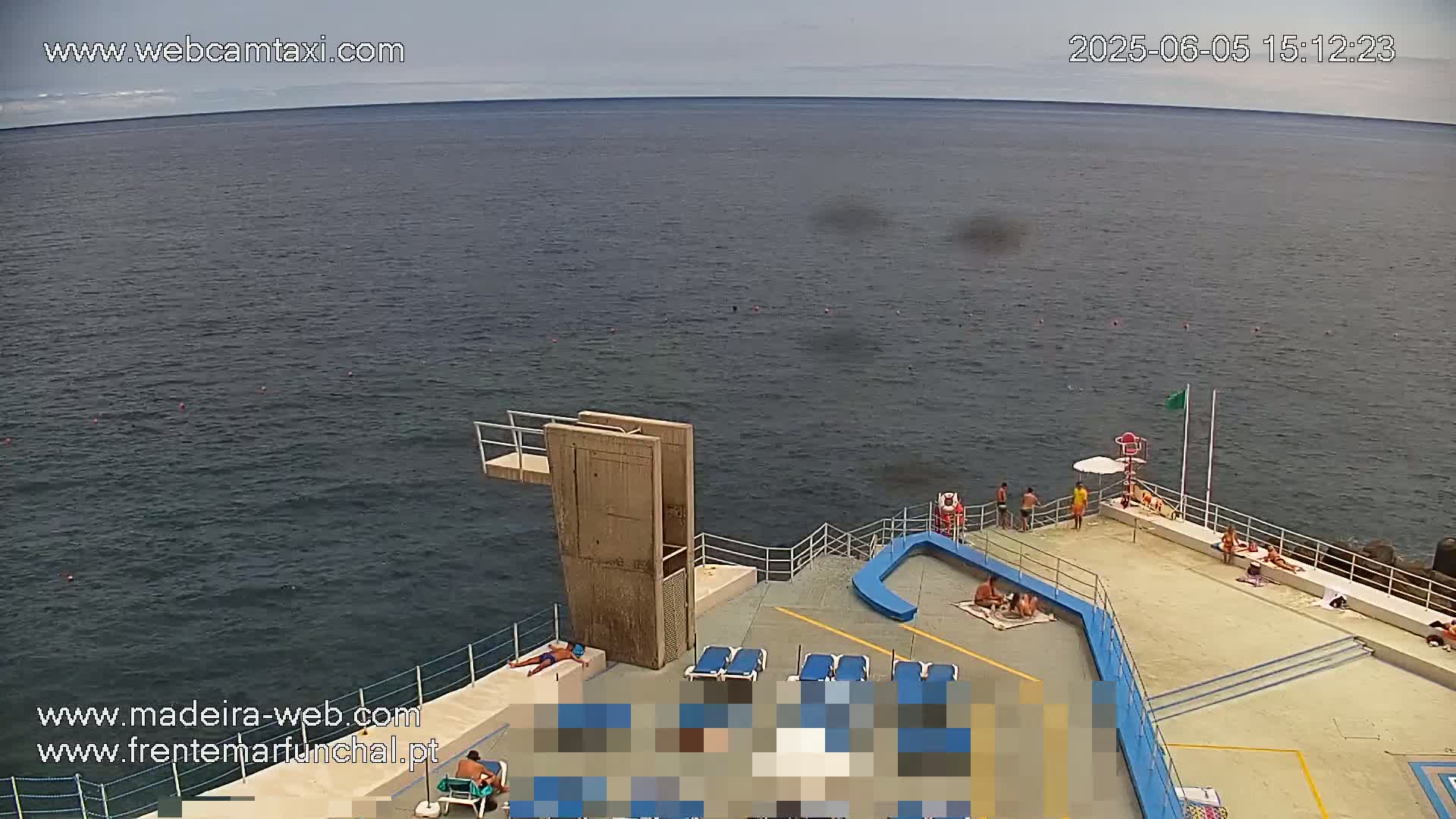 An overcast day at an outdoor pool area next to the ocean shows people sunbathing and swimming near a diving board.