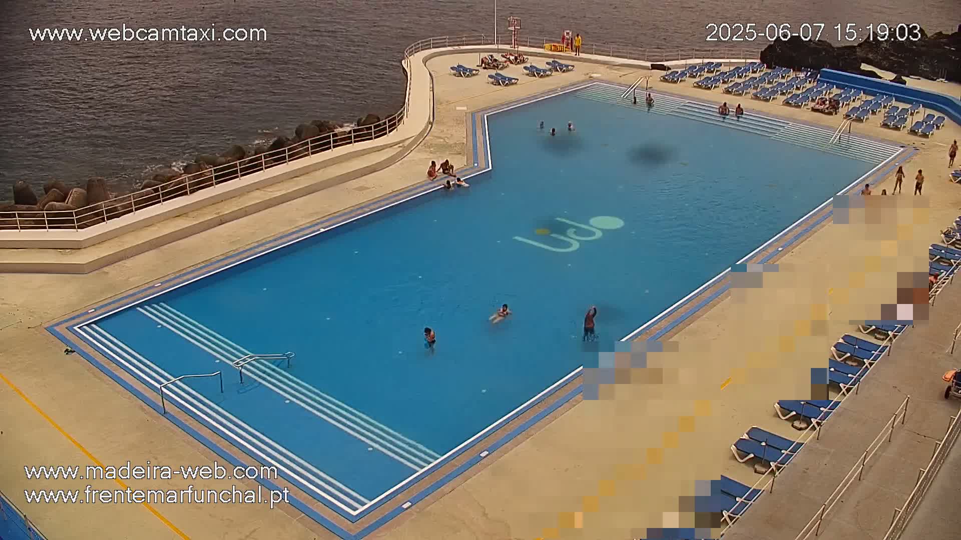 An outdoor swimming pool with several people swimming and sunbathing on surrounding deck chairs, next to a rocky ocean shore.