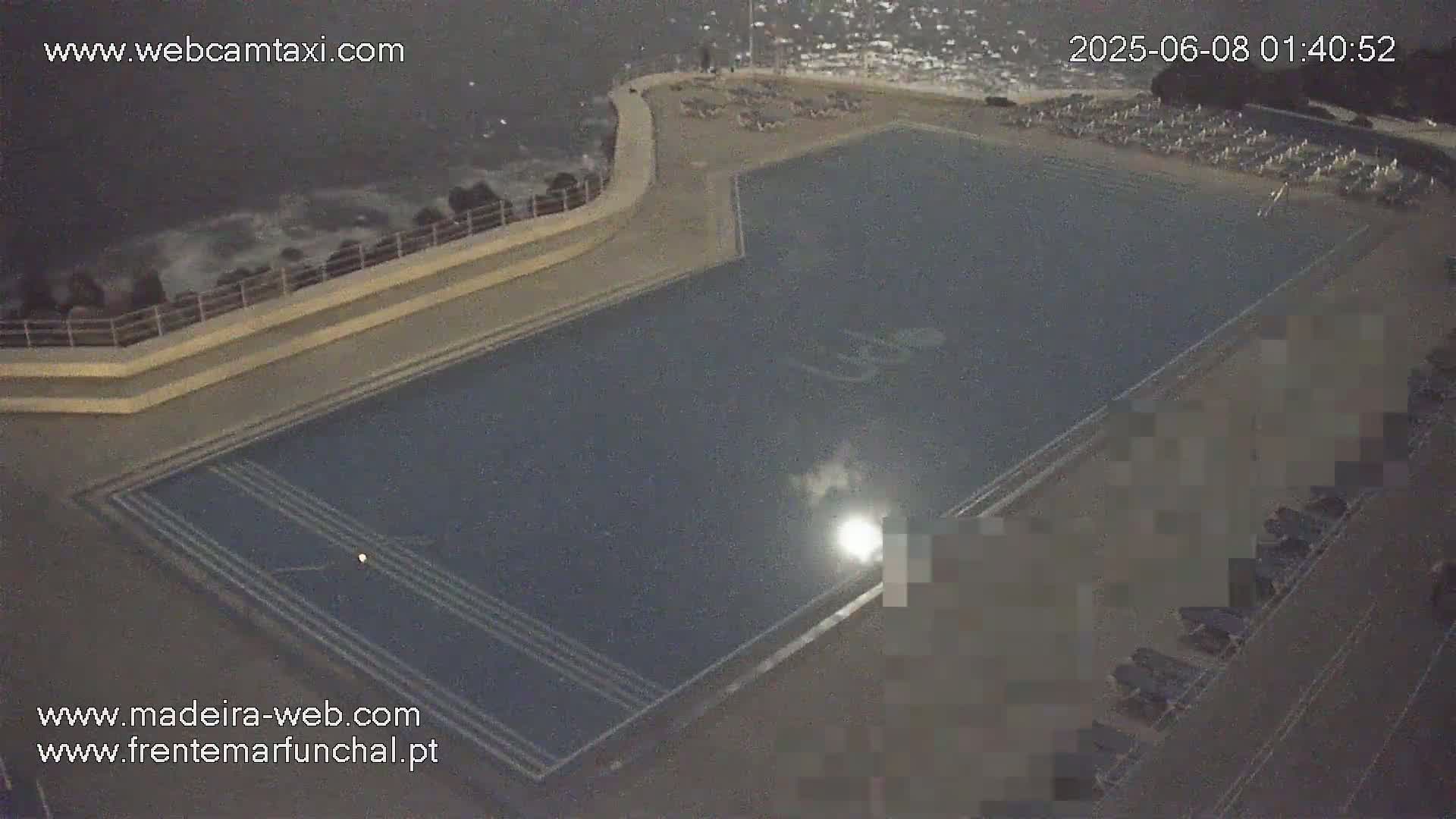 A large, dark blue outdoor pool sits beside a rocky coastline under a nighttime sky with choppy waves.
