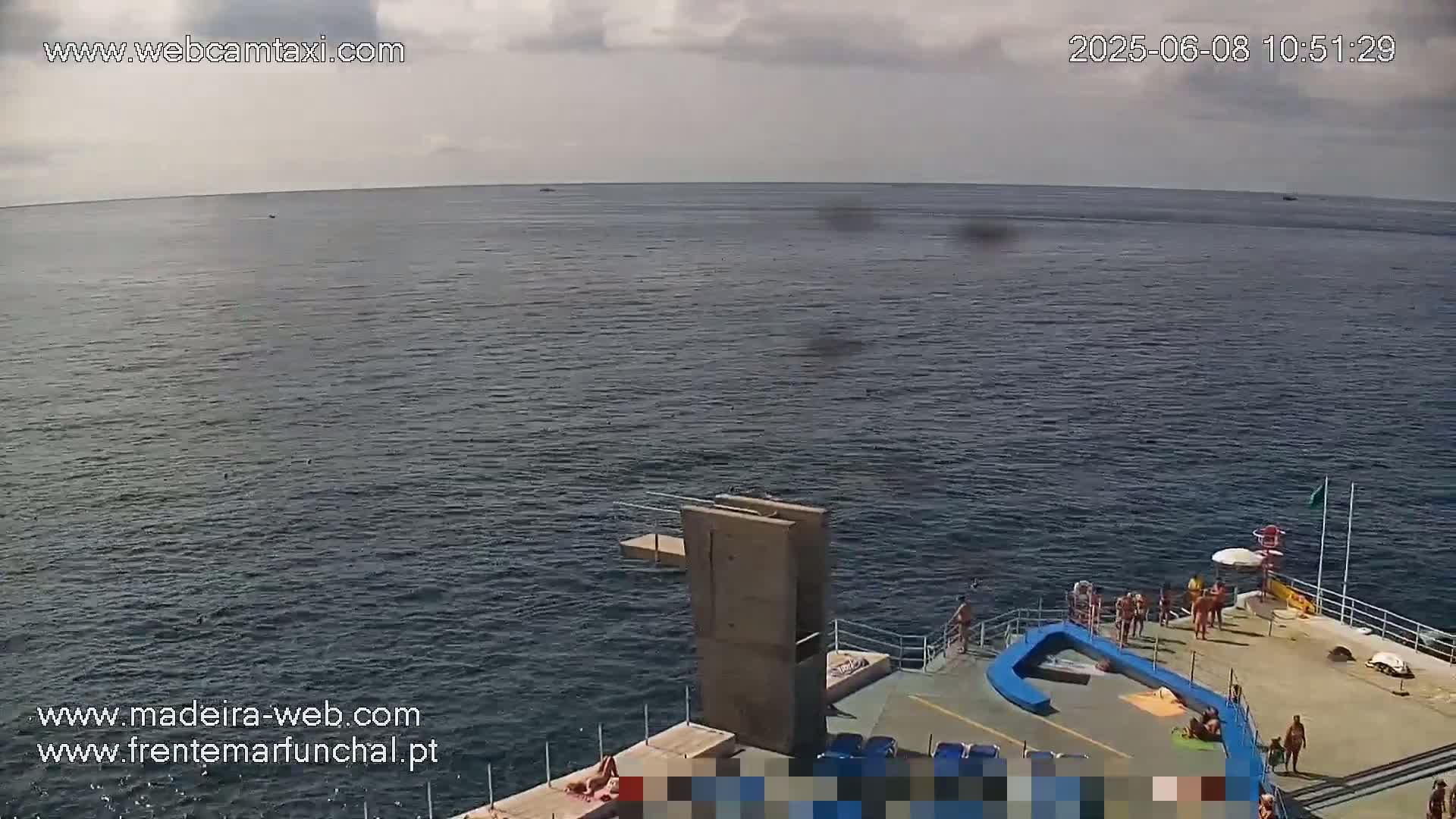 A mostly cloudy day shows a view of a calm ocean with a diving platform and several people sunbathing and swimming on a concrete platform next to it.