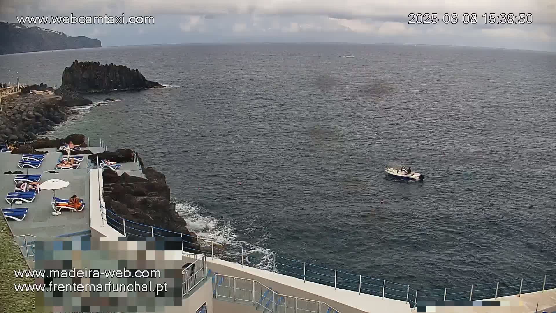 An outdoor scene shows a rocky coastline with a small, sunbathers-filled patio overlooking a dark-blue ocean where a small motorboat is visible under an overcast sky.