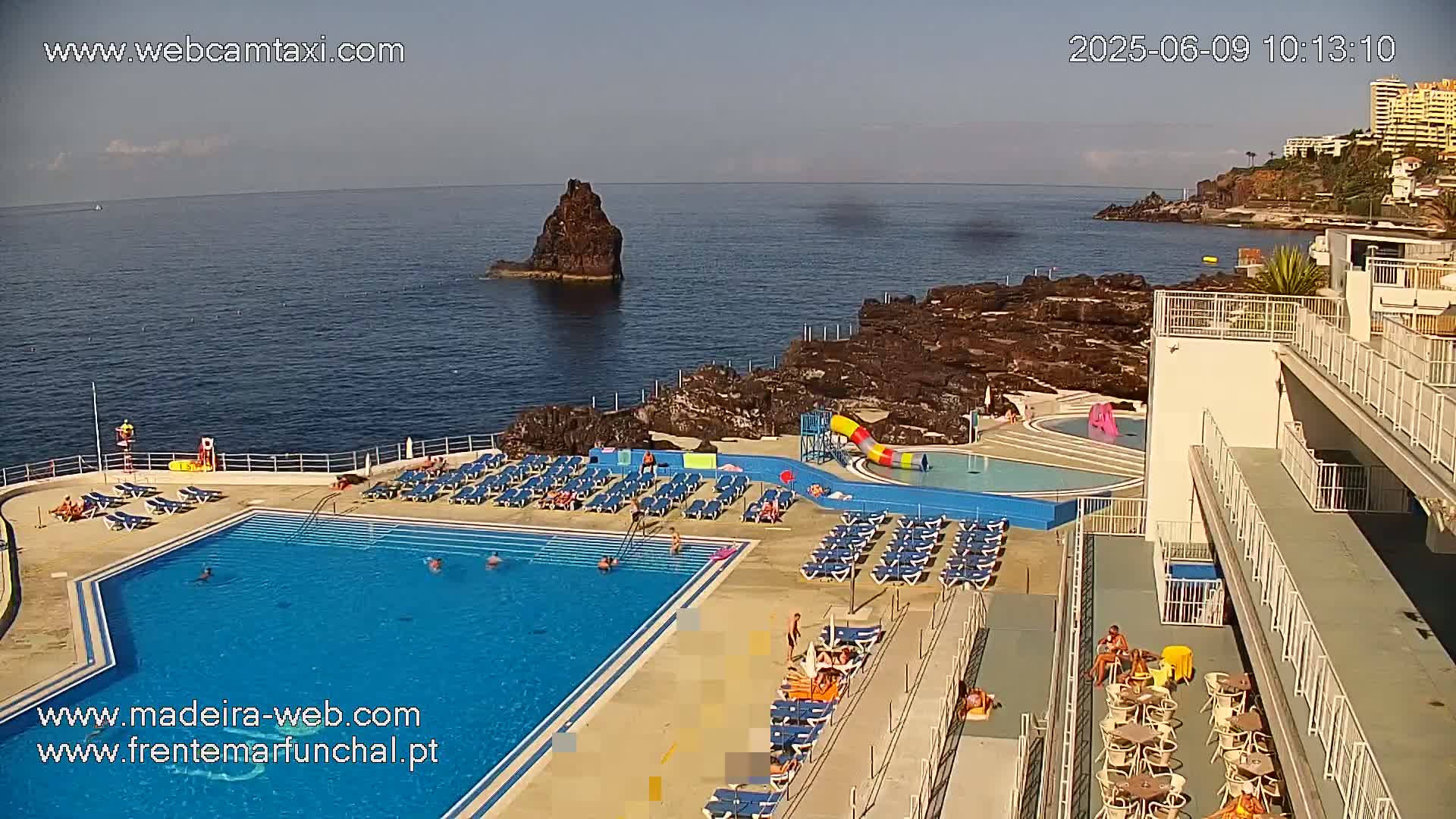 An outdoor pool and lounge area sits on a rocky coastline overlooking a calm ocean on a sunny day.