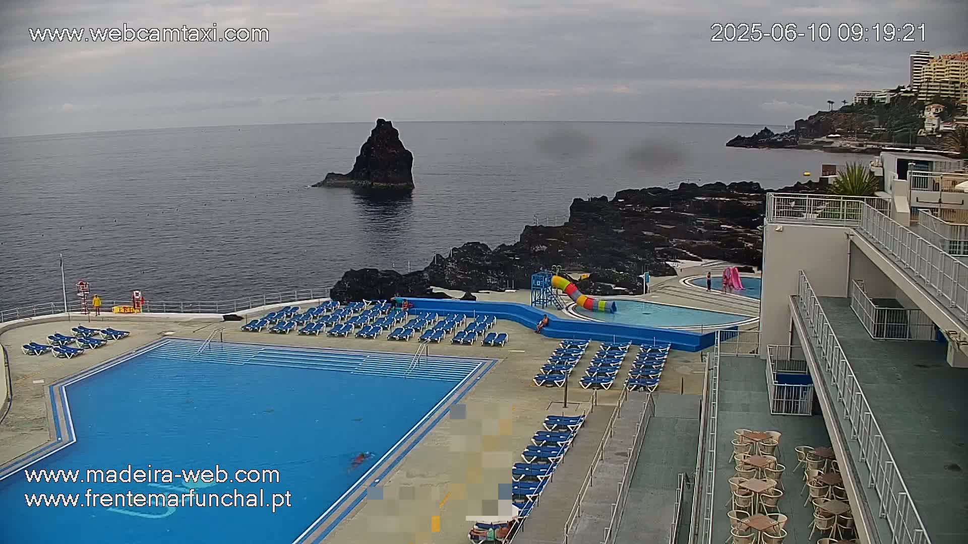 An outdoor pool and surrounding deck area overlook a dark-colored rocky coastline and ocean under an overcast sky.
