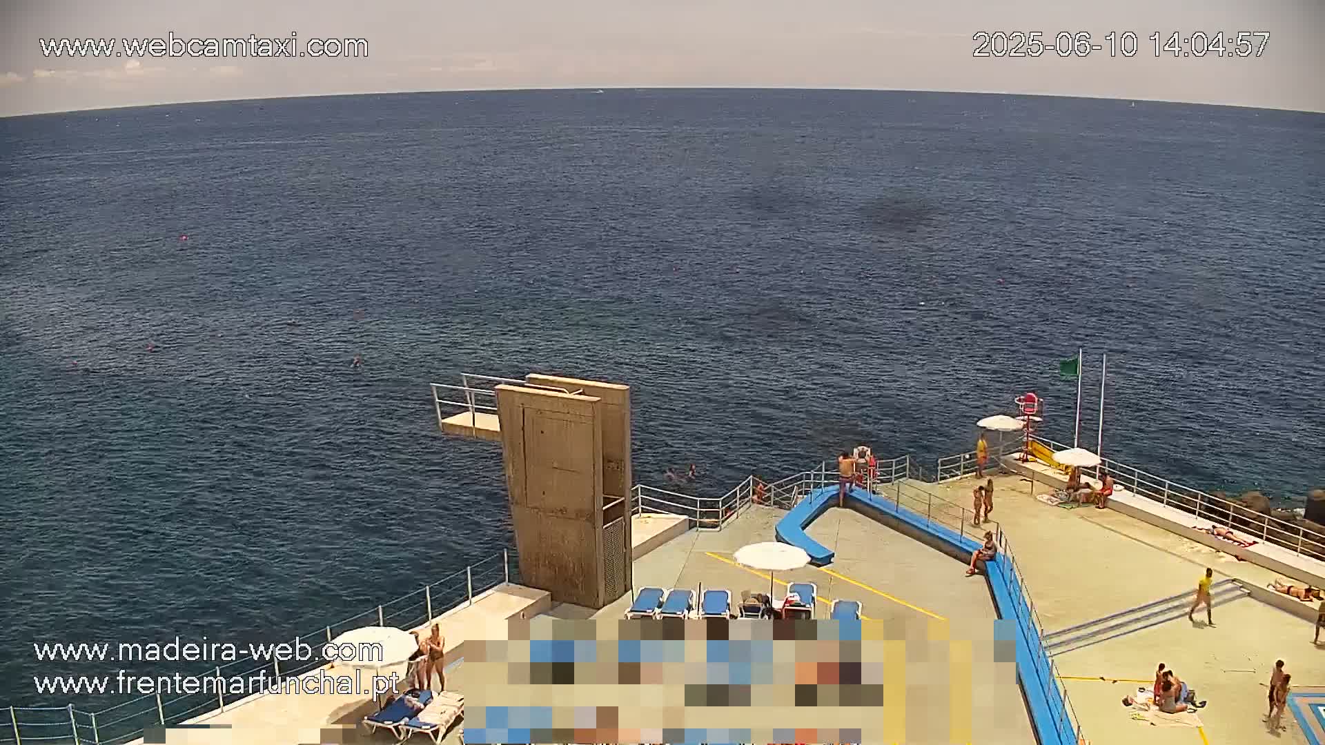 A high-angle view shows a seaside area with a diving platform, sunbathers, and swimmers on a sunny day.