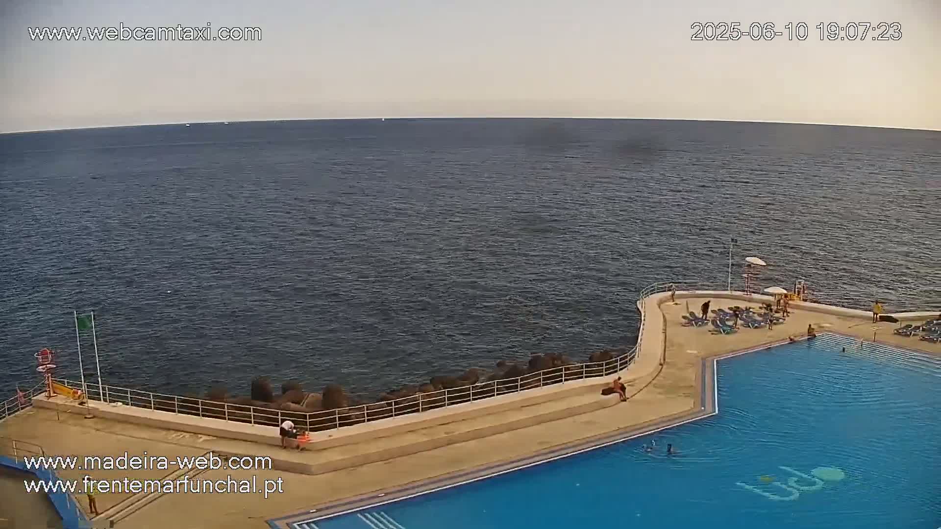 An outdoor pool complex with people swimming and lounging on the deck overlooks a calm ocean under a mostly clear sky.