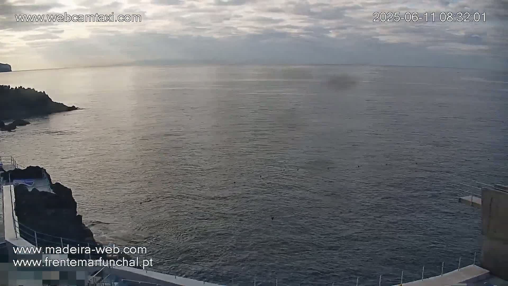 A calm ocean under a mostly cloudy sky is seen from an elevated vantage point, showing dark volcanic rock formations and a diving platform near the water's edge.