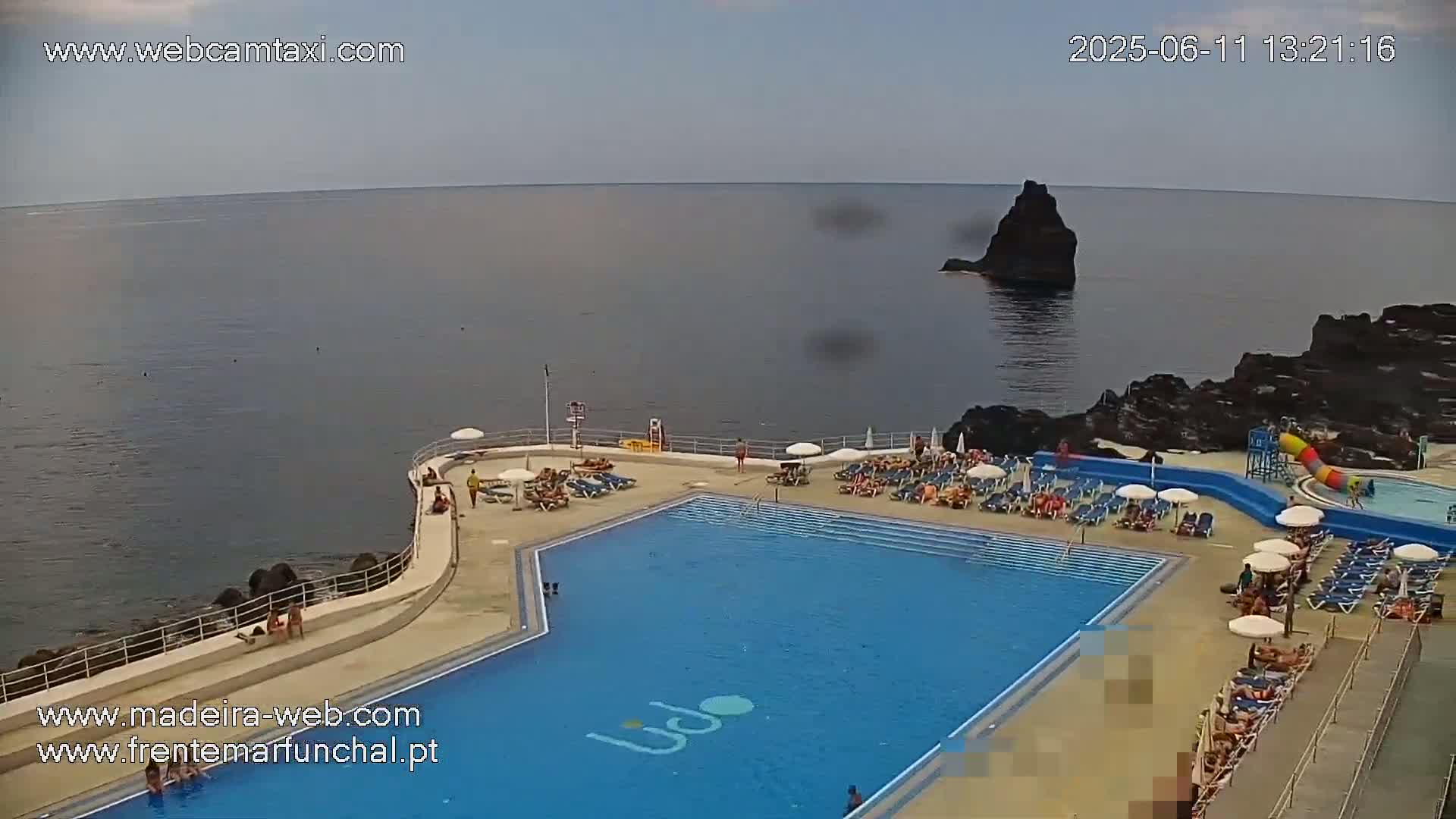 An outdoor pool with many sunbathers is situated on a rocky coast overlooking a calm ocean.