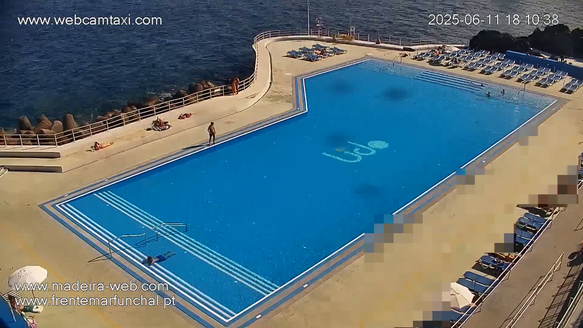 A large, rectangular outdoor pool with several people swimming and sunbathing on the surrounding deck on a sunny day next to the ocean.