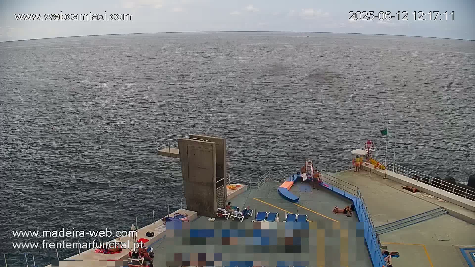 An overcast day shows a mostly calm body of water with a concrete diving platform and a sunbathing area with several people lounging on deck chairs.