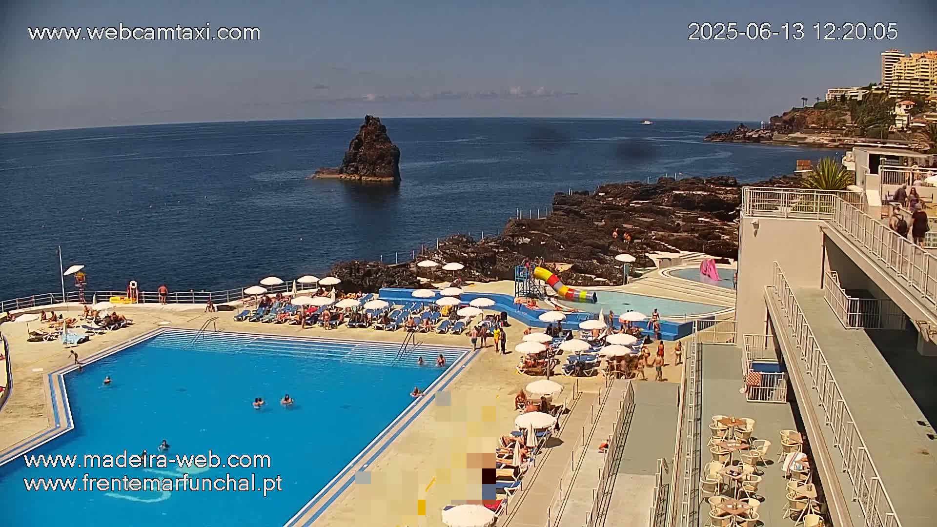 An outdoor pool complex with many people relaxing on lounge chairs and in the water, overlooking a rocky coastline and ocean on a sunny day.