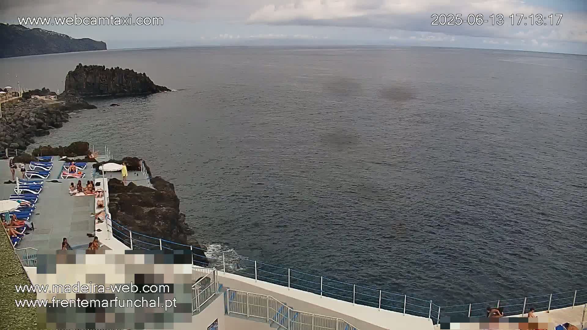 An outdoor scene shows a rocky coastline with a pool deck and people sunbathing, overlooking a calm ocean under an overcast sky.