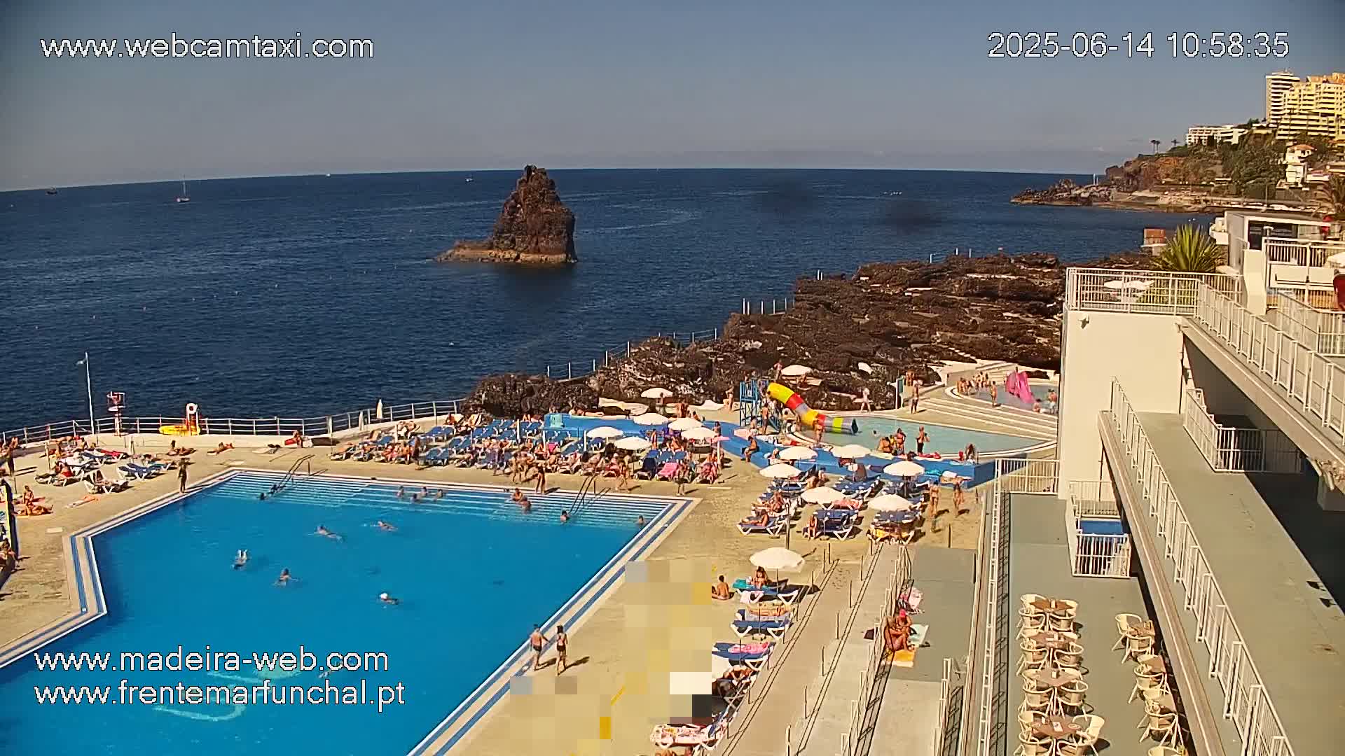 An outdoor pool area with many people relaxing and swimming, next to a rocky ocean coast on a sunny day.