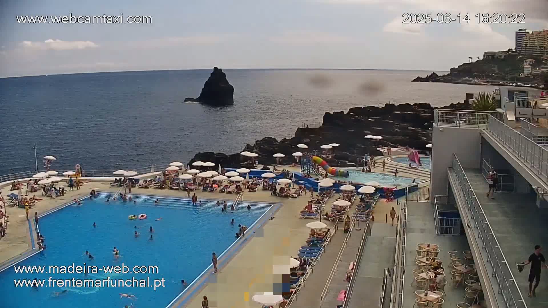 An outdoor pool area with many people swimming and lounging, adjacent to a rocky coastline and ocean under a mostly clear sky.