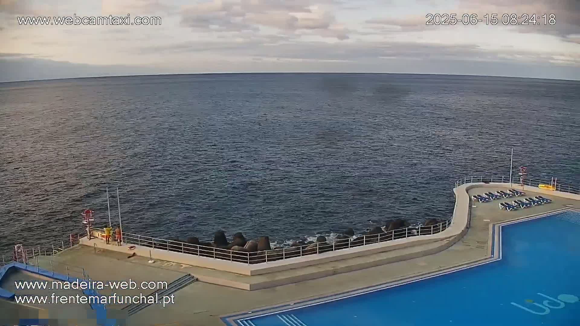 An outdoor swimming pool with lounge chairs sits next to a calm ocean under a mostly cloudy sky.