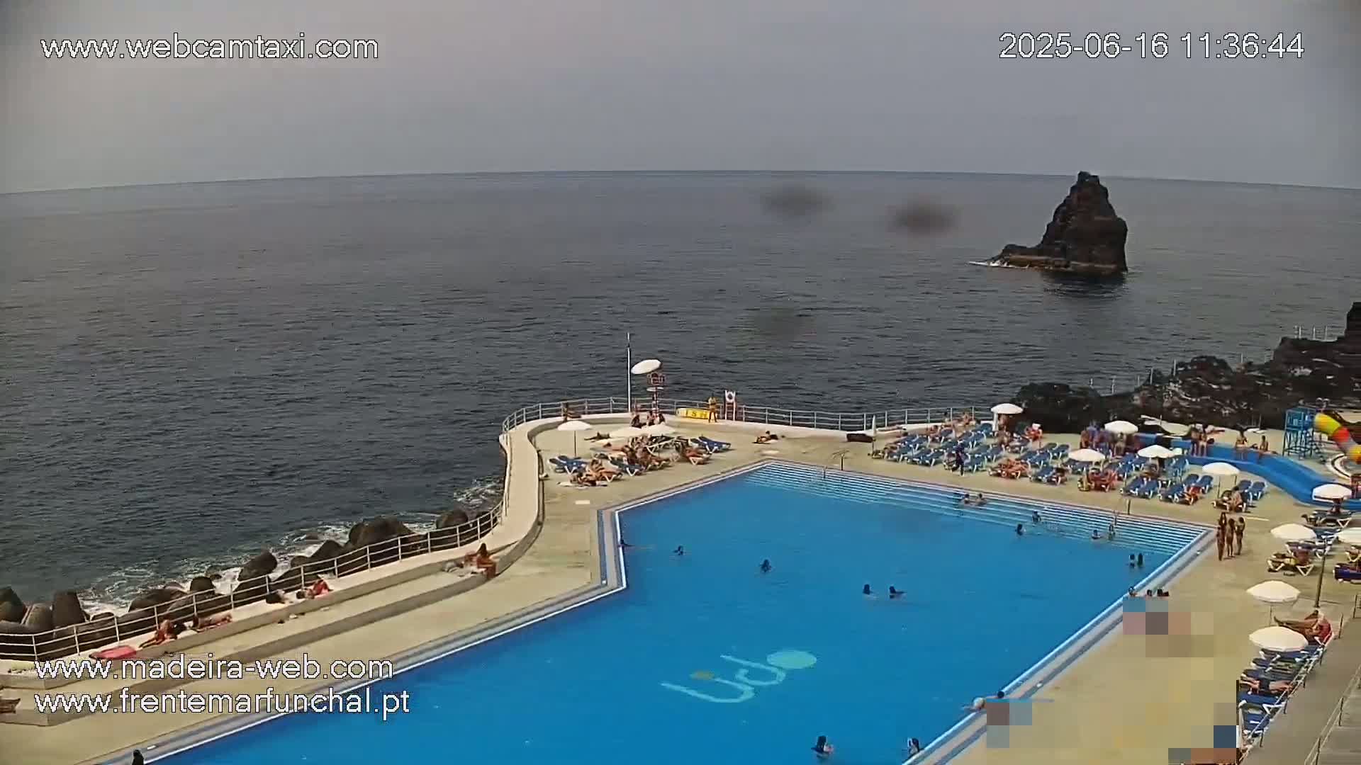 An outdoor swimming pool with many people swimming and sunbathing is situated on a rocky coast next to a dark-colored ocean under a mostly cloudy sky.