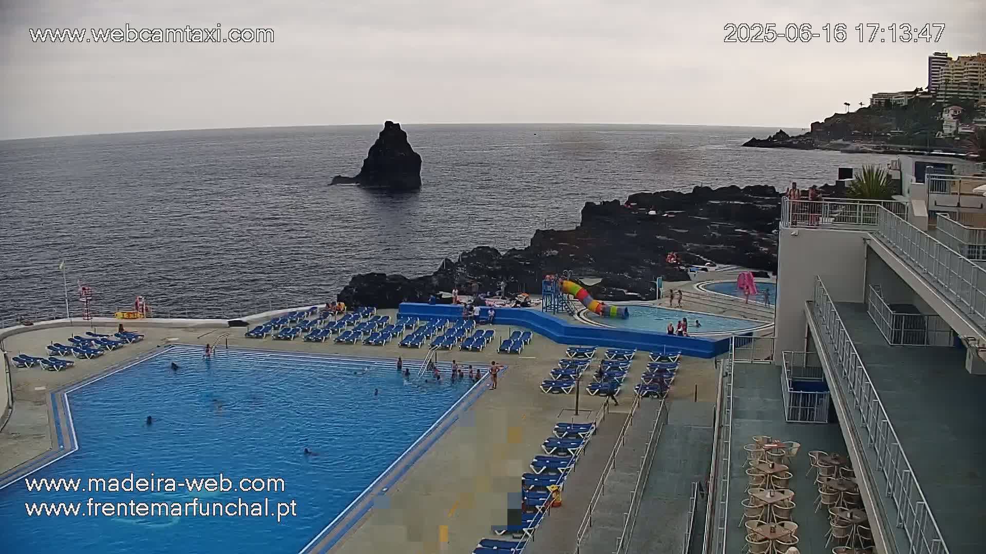 An outdoor pool complex with numerous lounge chairs and a water slide overlooks a dark-colored ocean under an overcast sky.