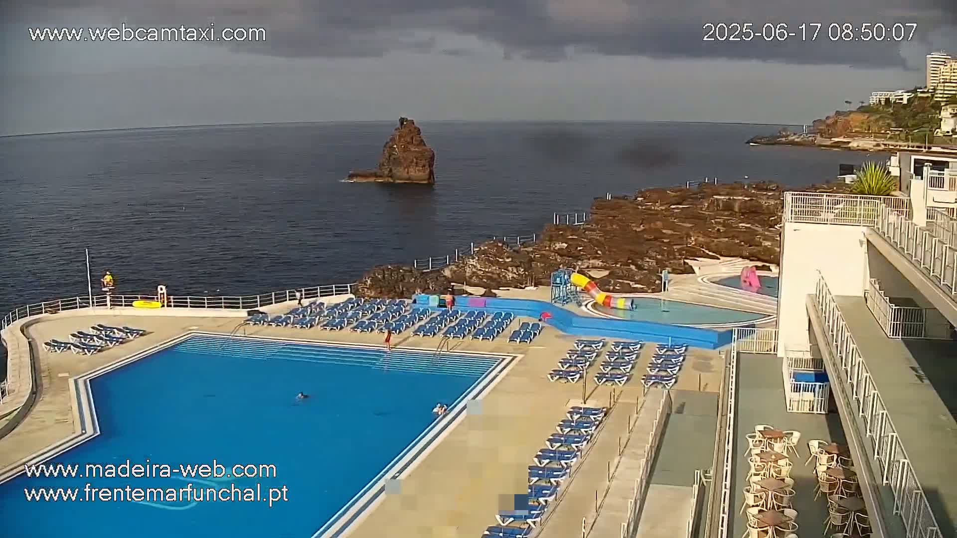 An outdoor pool area with lounge chairs surrounds a large, blue swimming pool next to a rocky coastline under partly cloudy skies.