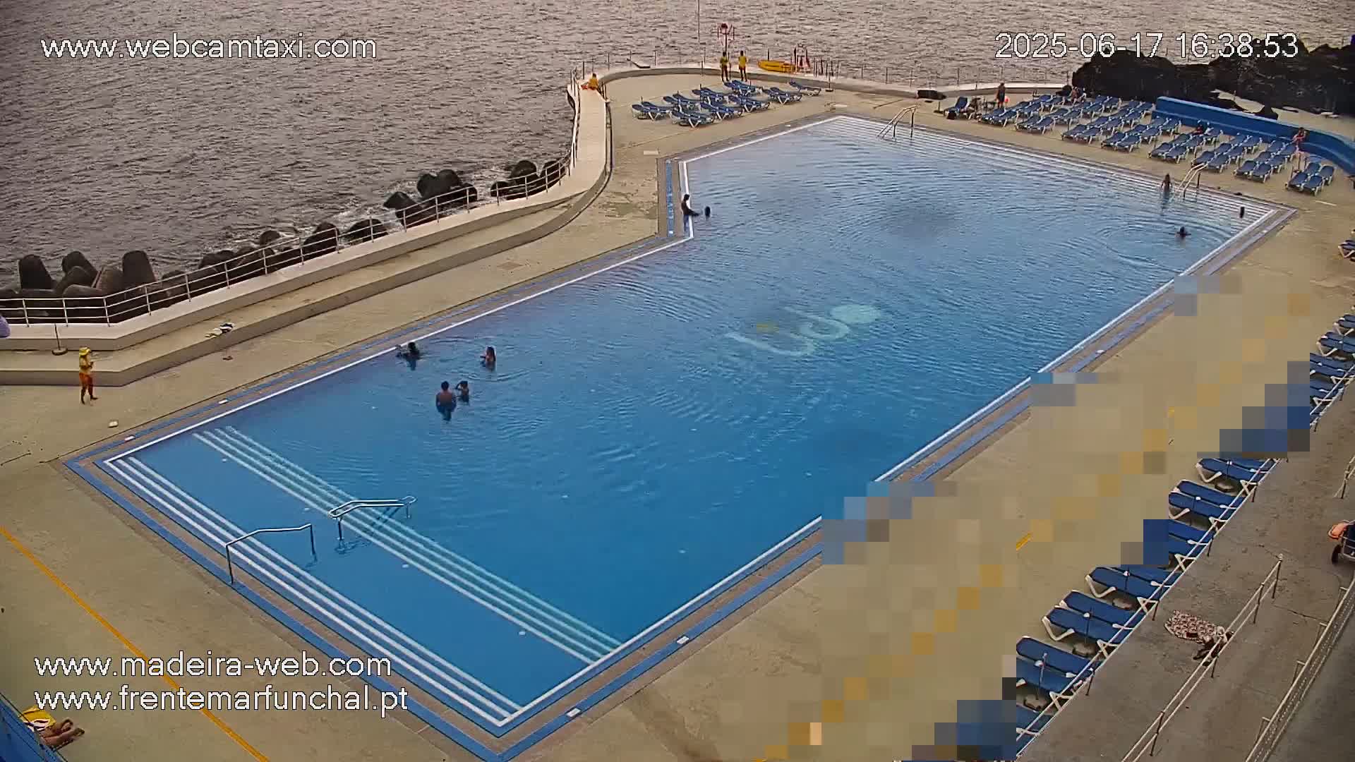 An outdoor swimming pool with several people swimming and sunbathing on the surrounding deck, next to a calm ocean.