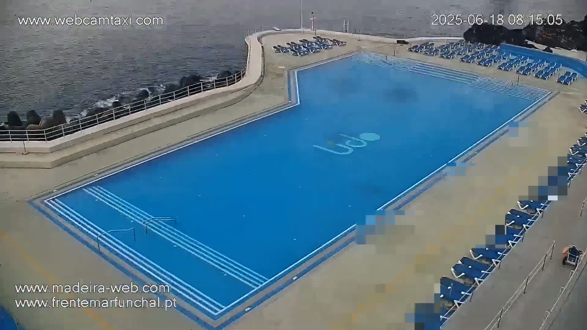 An outdoor, oceanside swimming pool with numerous empty lounge chairs sits beside a calm body of water.