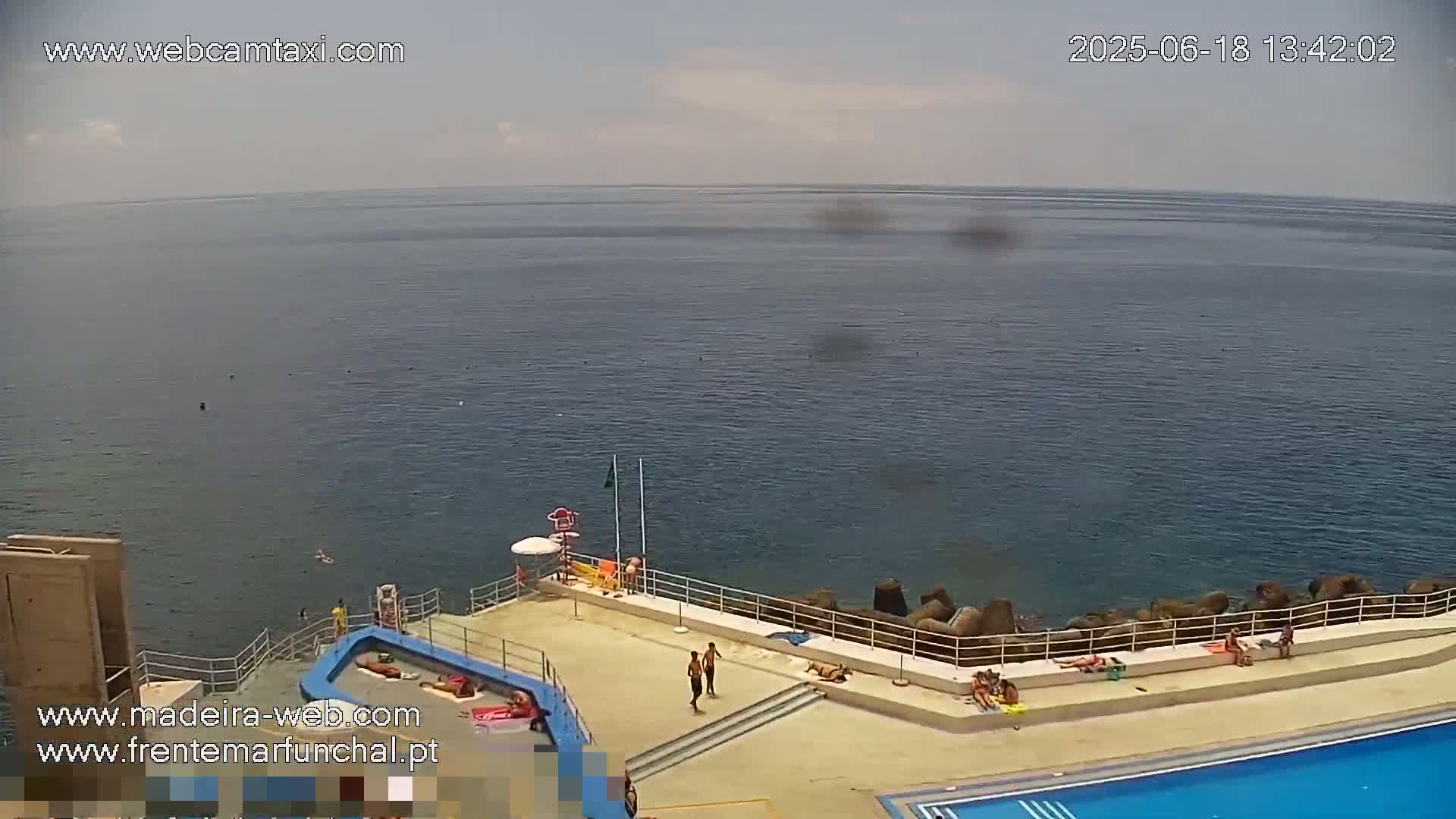 An outdoor pool area with sunbathers overlooks a calm ocean under a partly cloudy sky.