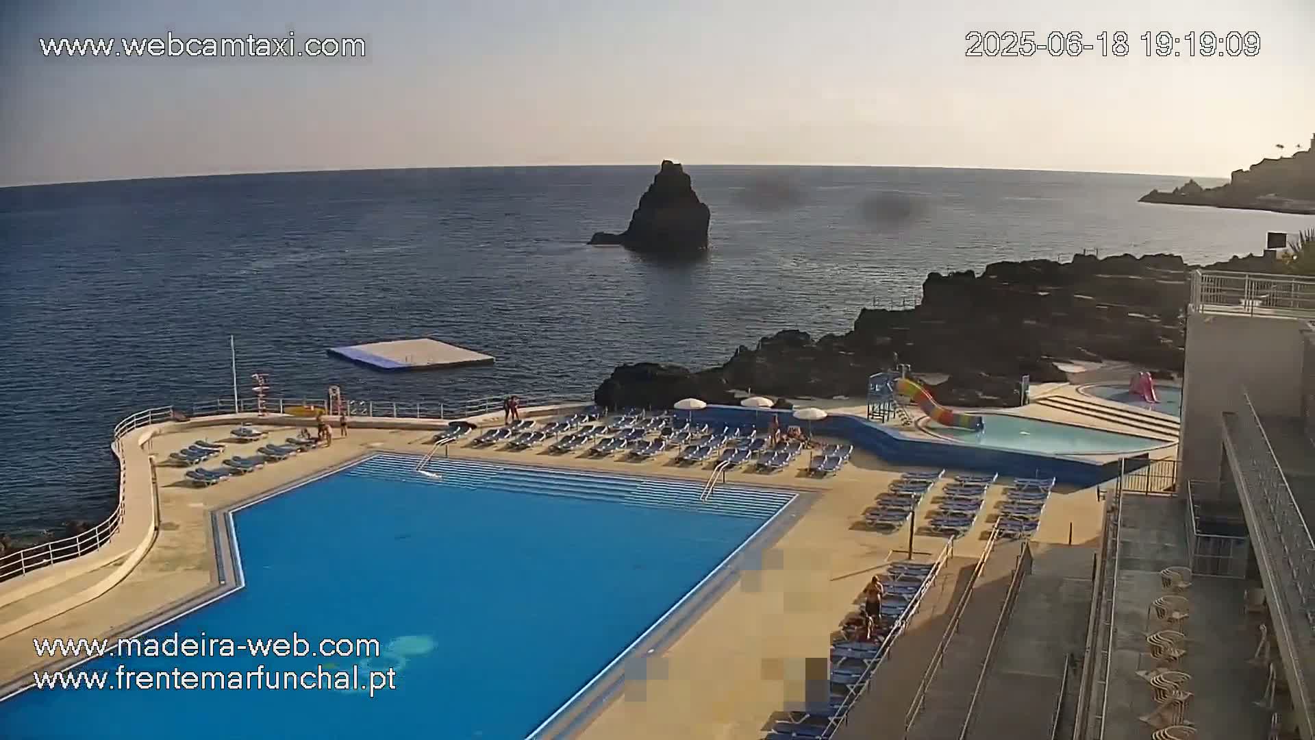 An outdoor pool with surrounding lounge chairs overlooks a calm ocean with a small island visible in the distance.