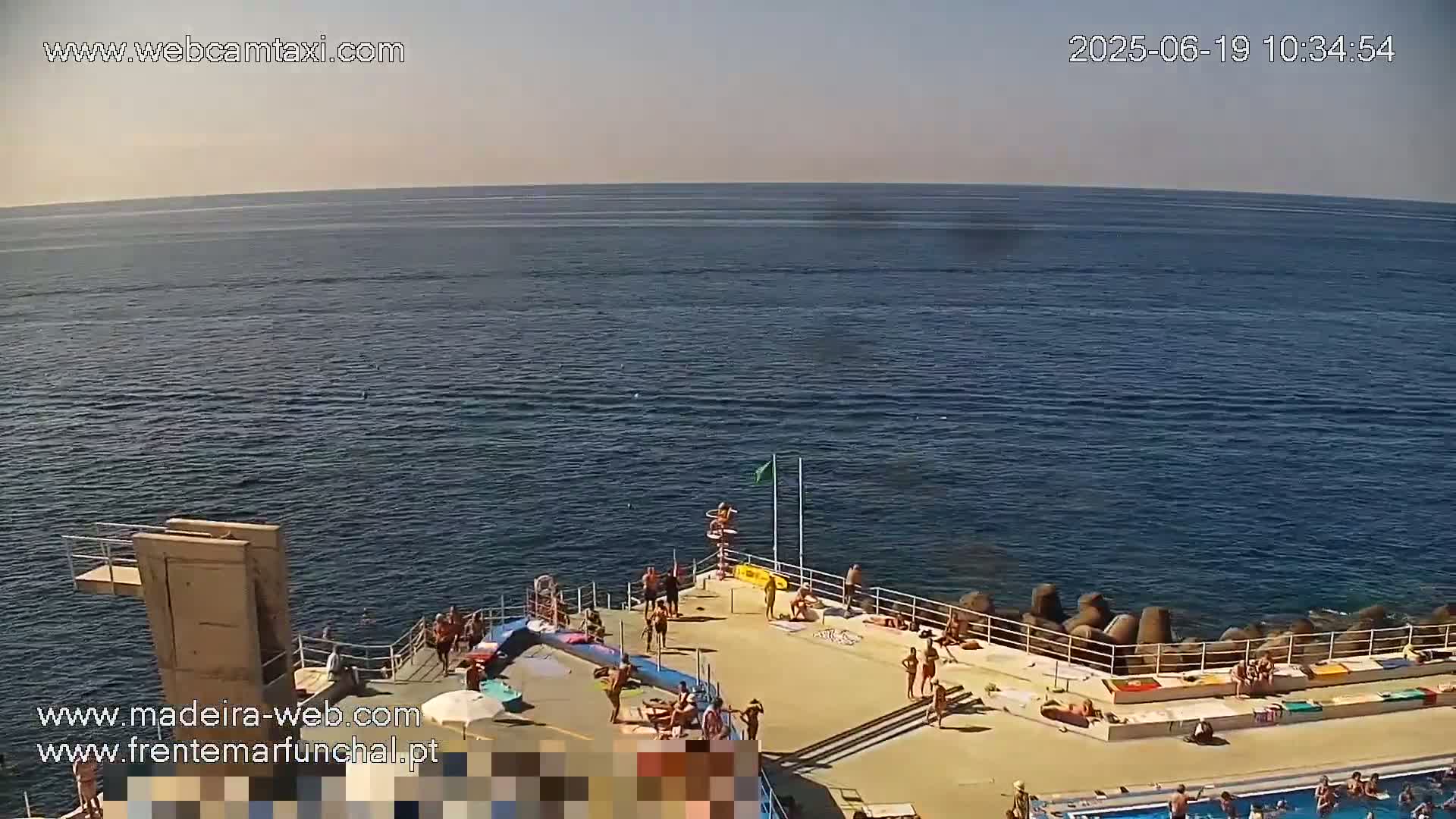 A sunny day at an outdoor pool area overlooking a calm ocean, with people swimming, sunbathing, and lounging around.
