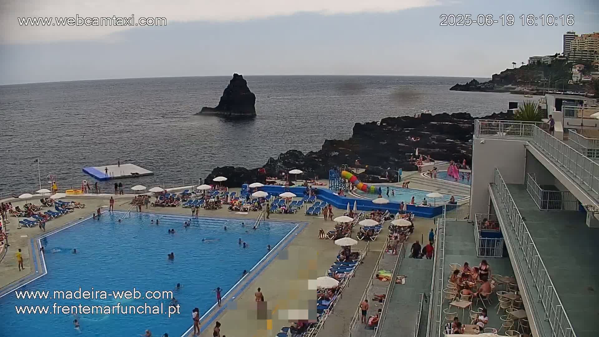 An outdoor pool complex with many people swimming and lounging, situated on a rocky coastline overlooking a calm ocean under a partly cloudy sky.