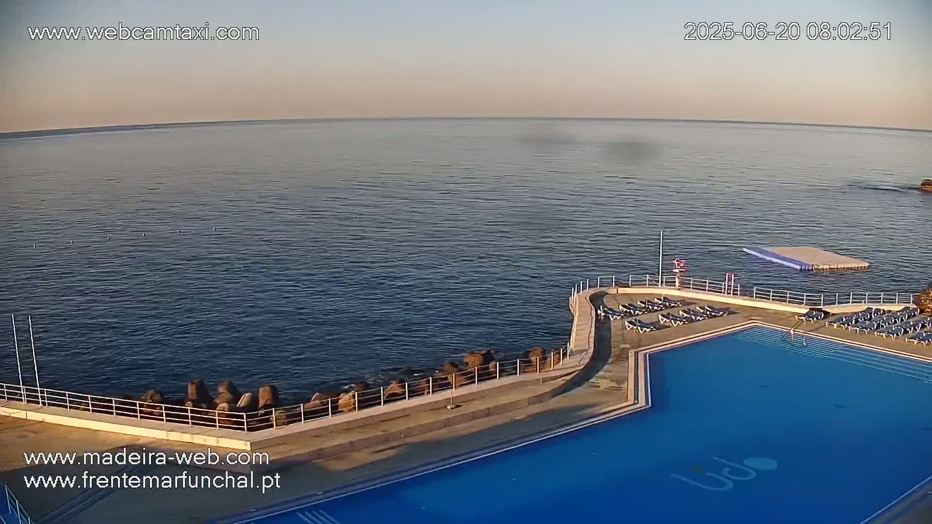An outdoor swimming pool with lounge chairs sits beside a calm ocean on a clear, bright morning.