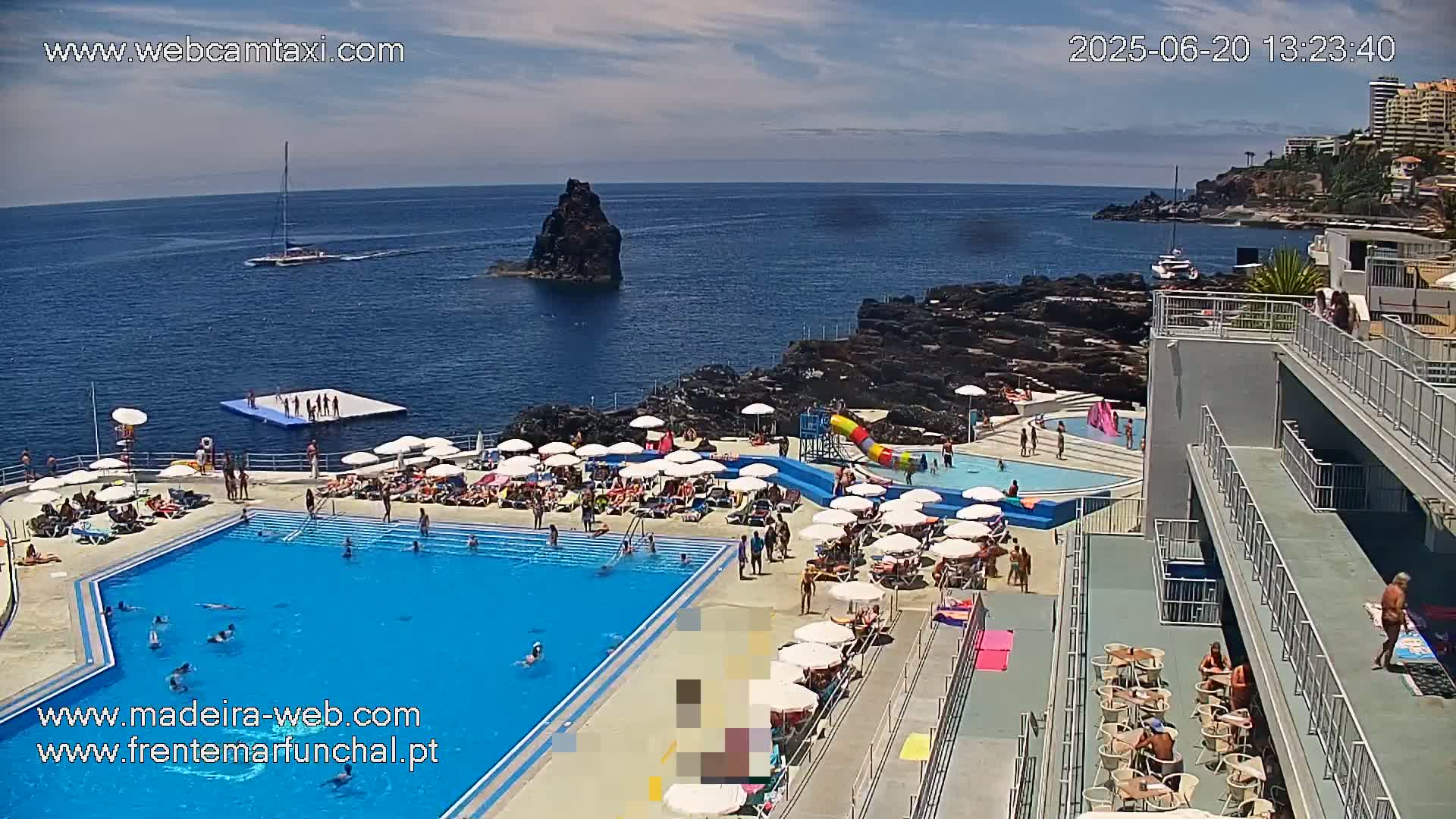 A large outdoor pool surrounded by sunbathers and lounge chairs is situated on a cliff overlooking a calm ocean with a small sailboat and rocky outcroppings under a partly cloudy sky.