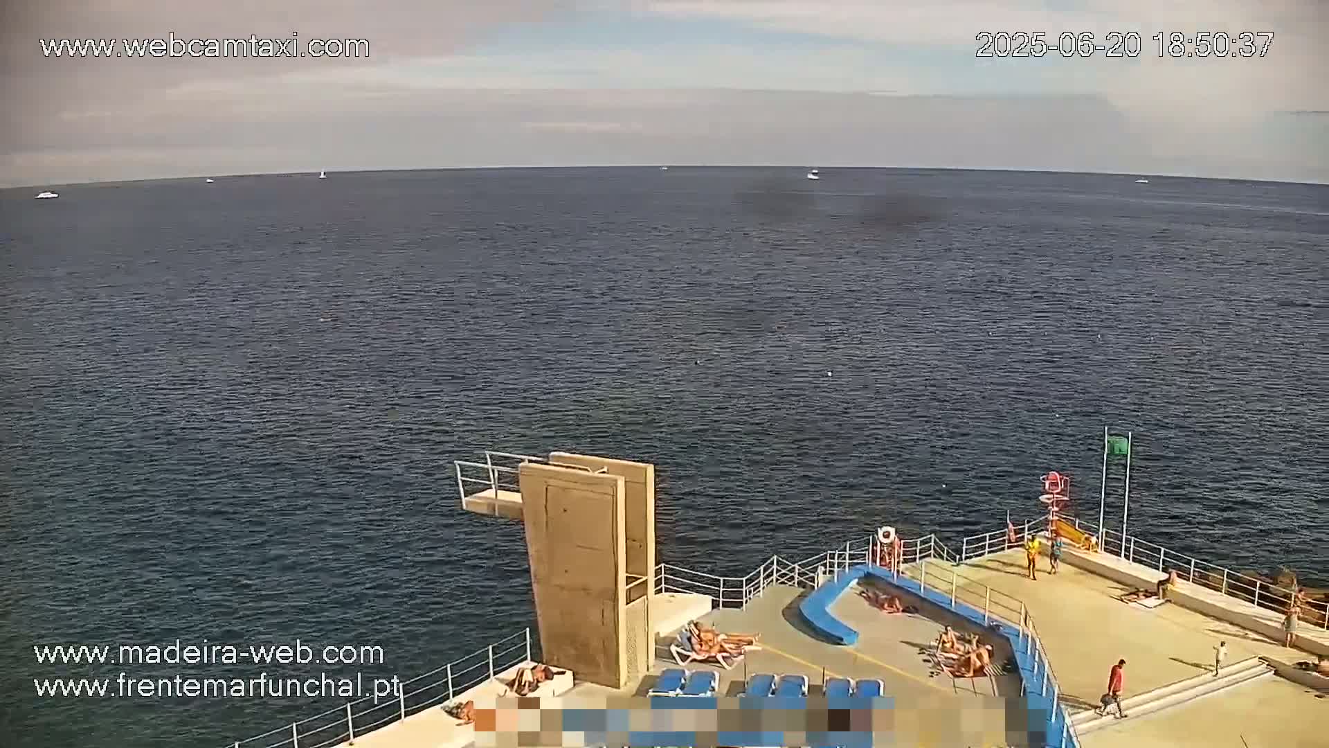 A mostly sunny day shows a calm ocean with a few boats in the distance, and a concrete diving platform with several people sunbathing and swimming near it.